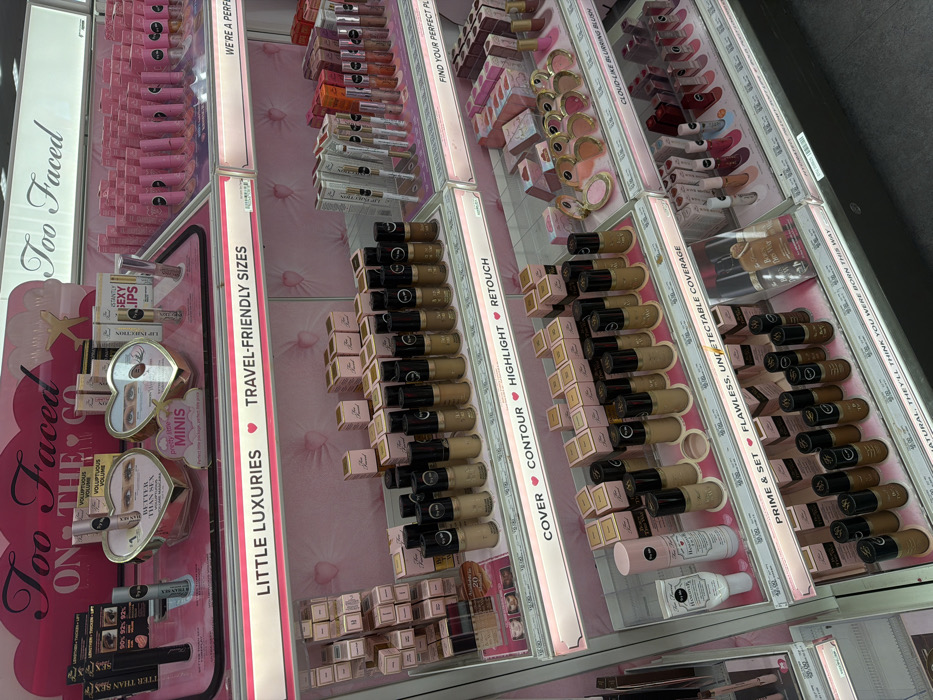 This image depicts a brightly illuminated retail cosmetics display for the "Too Faced" brand, located in Miami, United States of America. The multi-tiered display features clear acrylic shelves with pink, tufted back panels.
The top shelf showcases "Too Faced ON THE GO" products, including "Better Than Sex" mascara displayed within heart-shaped stands and tubes of "Lip Injection" lip plumper. Signage indicates "LITTLE LUXURIES" and "TRAVEL-FRIENDLY SIZES."
The middle and lower shelves are segmented by function, labeled "COVER," "CONTOUR," "HIGHLIGHT," "RETOUCH," "PRIME & SET," and "FLAWLESS, UNDETECTABLE COVERAGE." These sections contain numerous "Too Faced" foundation and concealer bottles in a range of skin tones, characterized by black caps and beige product bases, along with associated product boxes. Additionally, primers and setting sprays, such as "Hangover Rx," are present.
To the right, additional shelves display an assortment of "Too Faced" lip products, including glosses and lipsticks, alongside heart-shaped compacts identified as "CLOUD-LIKE BLURRING BLUSH." Price tags are visible beneath various items. The flooring at the base of the display is dark gray. No individuals are present in the image.