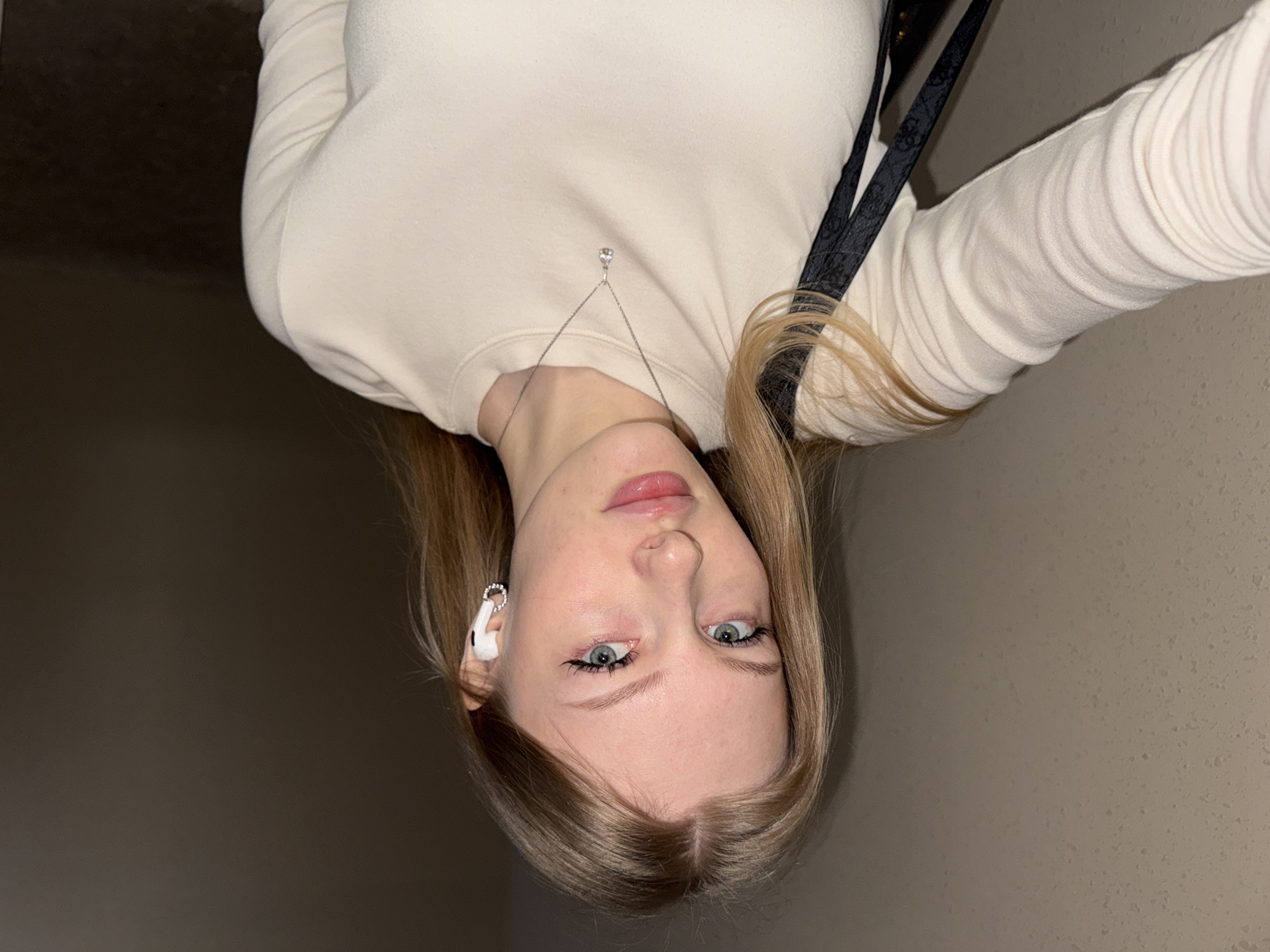 A selfie taken by a young Caucasian woman with long blonde hair and blue eyes, wearing a light-colored long-sleeved top and a delicate necklace with a small pendant. She is wearing a white earpiece in her left ear. The background appears to be an interior wall, possibly in a building in Perm, Russia, as indicated by the user's prompt. The photo is taken at an angle, with the camera positioned above her.