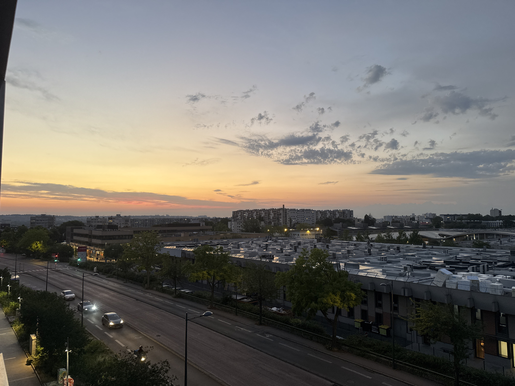 The image is a high-angle, wide shot of a city street at dusk. The sky is a gradient of orange and yellow near the horizon, fading into a pale blue and then gray towards the top, with scattered clouds. On the left side of the image, there is a multi-lane road with several cars driving, one of which is a motorcycle with a rider. Streetlights and traffic lights are visible along the road. Trees line the side of the road, some of which are illuminated. In the background, there are buildings, including a large, flat-roofed industrial or commercial building covered in what appear to be air conditioning units or vents. Further in the distance, a cluster of taller residential buildings are visible. The scene suggests an urban environment during the transition from daylight to night.