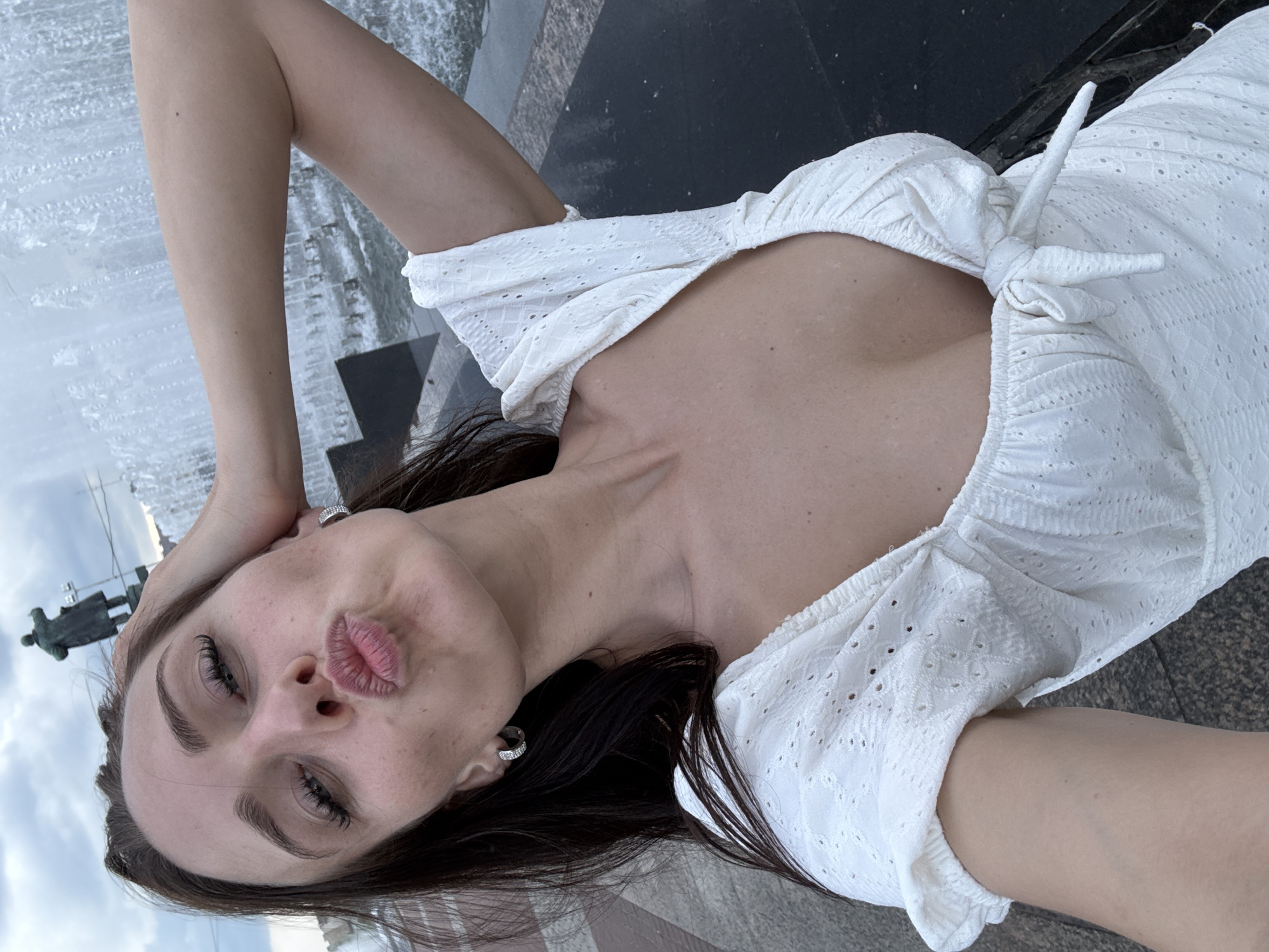 The image shows a woman posing for a selfie in front of a fountain. She is wearing a white dress and has her arm raised. Behind her, a statue is visible in the background. The fountain appears to be part of a monument or public square. The woman is pouting her lips, and her hair is dark brown. The sky is cloudy. The statue in the background is of a man in a long coat, possibly a historical figure. The fountain is spraying water upwards, creating a misty effect. The paving stones in the foreground suggest an outdoor public space. The overall scene is typical of a tourist attraction in a city.
