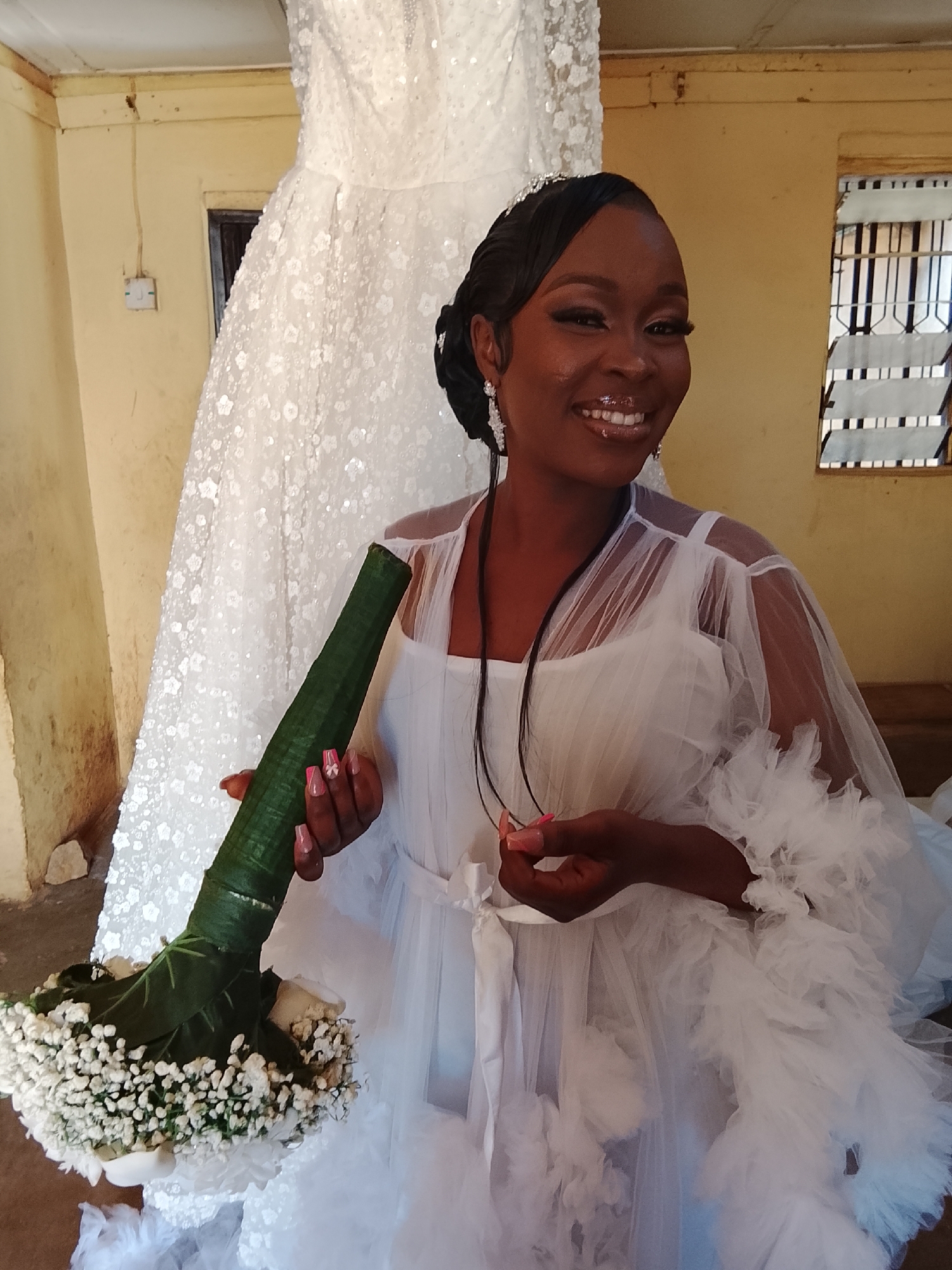 In an indoor setting in Unknown, Unknown, a dark-skinned woman is smiling at the camera, holding a wedding bouquet of white flowers wrapped in a green covering. She is wearing a white, sheer robe over a white garment. Behind her is a full wedding gown with a sequined pattern, and the walls are painted a light yellow. A window with bars is visible in the background. A light switch is mounted on the wall.
