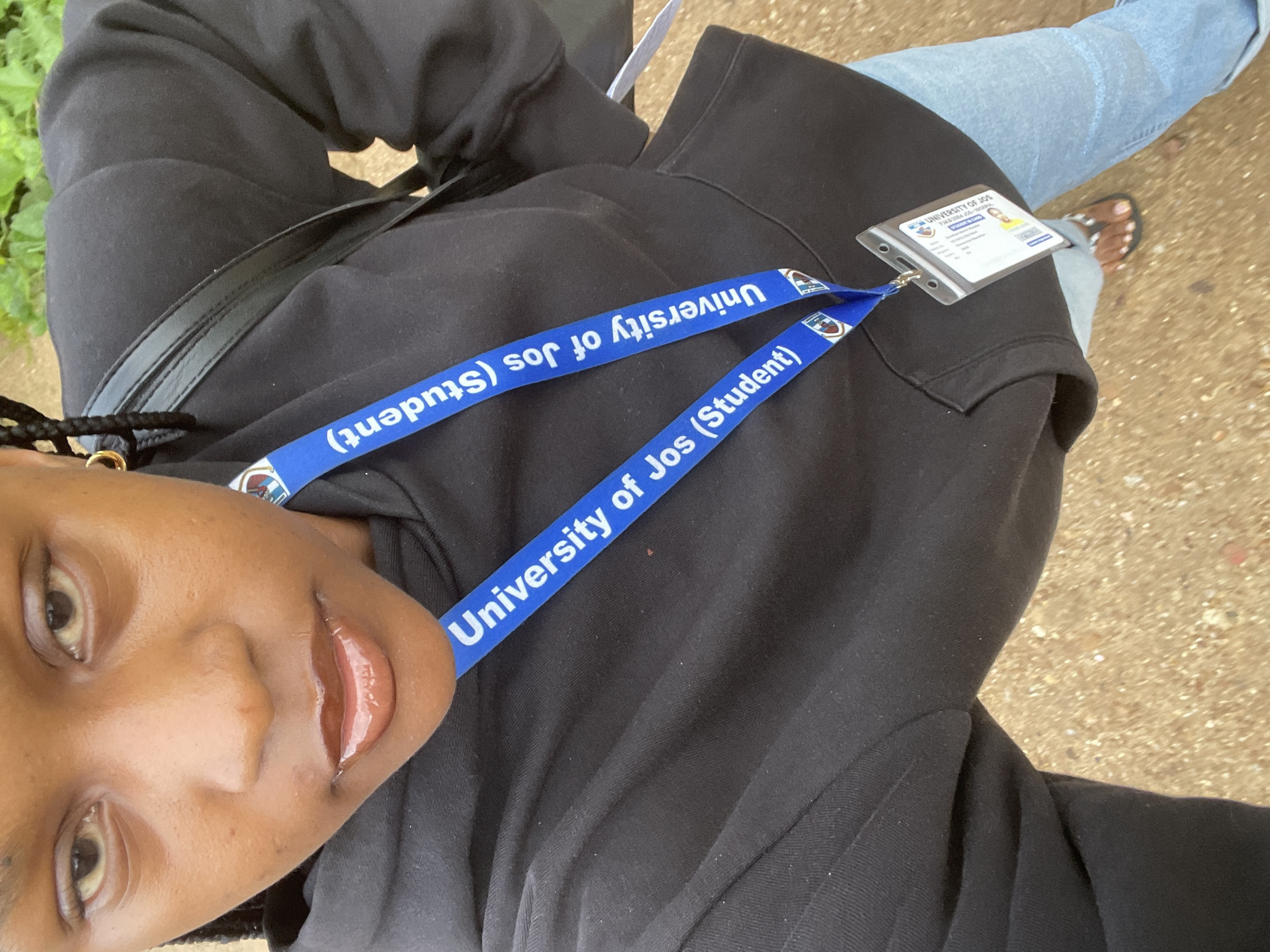 The image is a close-up, high-angle selfie of a young Black person wearing a black hoodie and light blue jeans. A black strap of a bag is visible across the hoodie. Attached to the strap are two blue lanyards with the text "University of Jos (Student)" printed in white, along with the university's crest. One lanyard has a clear plastic holder containing an ID card. The ID card is for the "UNIVERSITY OF JOS" and identifies the holder as a "STUDENT ID CARD" with fields for "Surname," "First Name," "Matric No.," "Faculty," "Department," and "Session." A small photo of the student is on the ID.
The person's face is the most prominent subject, with their eyes looking towards the camera and their lips slightly parted, appearing to have a glossy sheen. Their skin tone is dark, and their hair is braided and partially visible. The background is out of focus, showing glimpses of green foliage and a sandy or gravelly surface.
The overall impression is casual and personal, likely taken by the student themselves on the university campus or in a nearby area in Rafin Par, Nigeria. The presence of the student ID and lanyards clearly indicates an educational setting and the student's affiliation with the University of Jos. The lighting suggests it is daytime, and the clothing implies a warm climate.