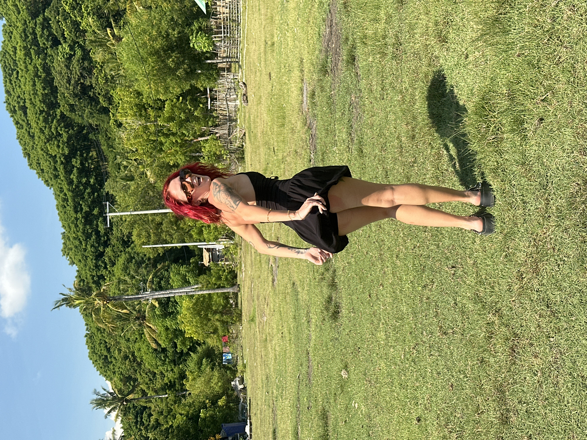 A woman with red hair and sunglasses is standing in a grassy field in Unknown, Indonesia. She is wearing a short black dress and black shoes. The background includes trees and a blue sky.