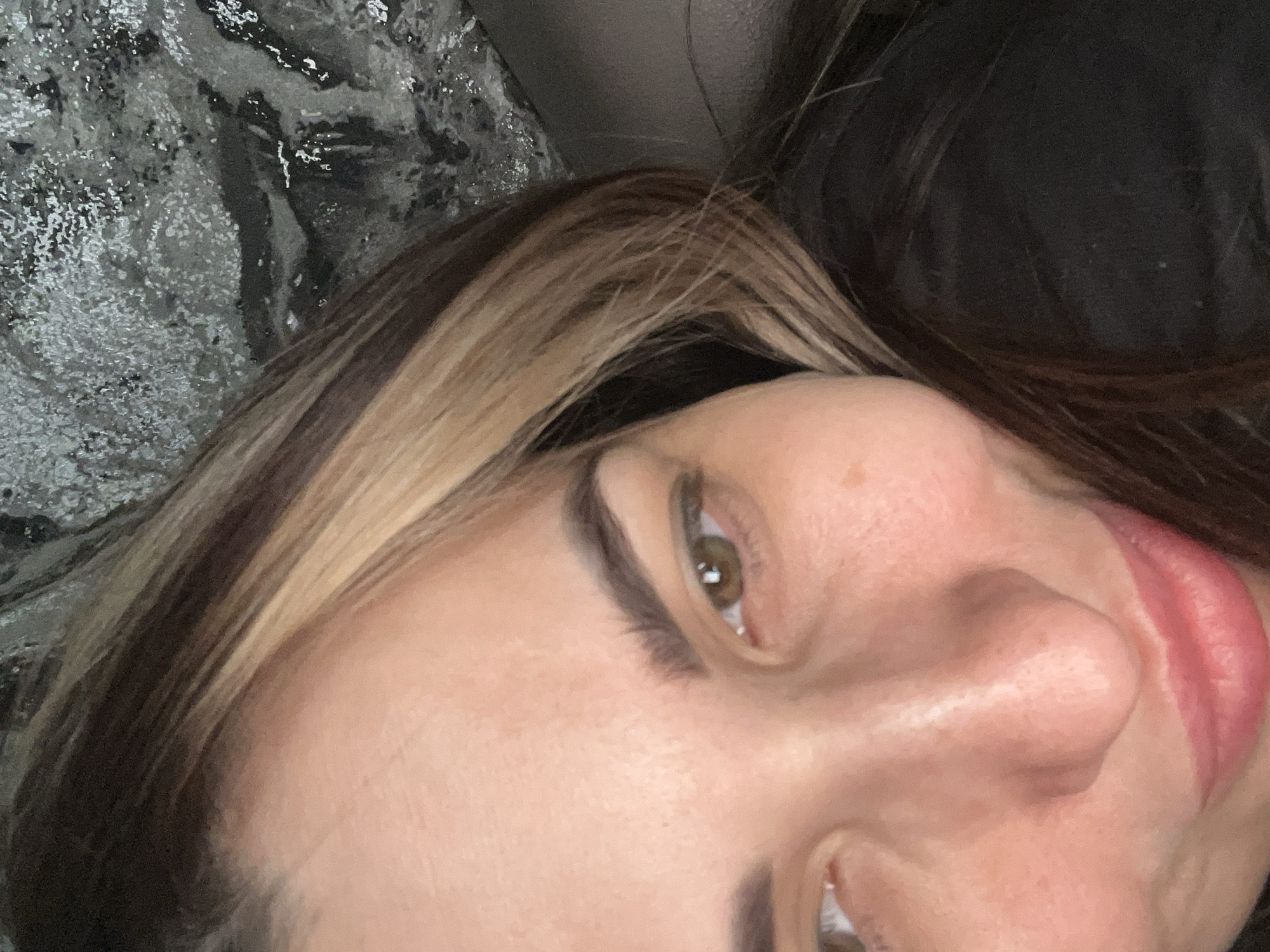 A close-up, slightly angled portrait of a person with fair skin, brown hair with blonde highlights, and hazel eyes. The person's nose, lips, and eyebrows are visible. The background includes a textured gray material and a dark-colored soft material. The location is Unknown, Unknown.