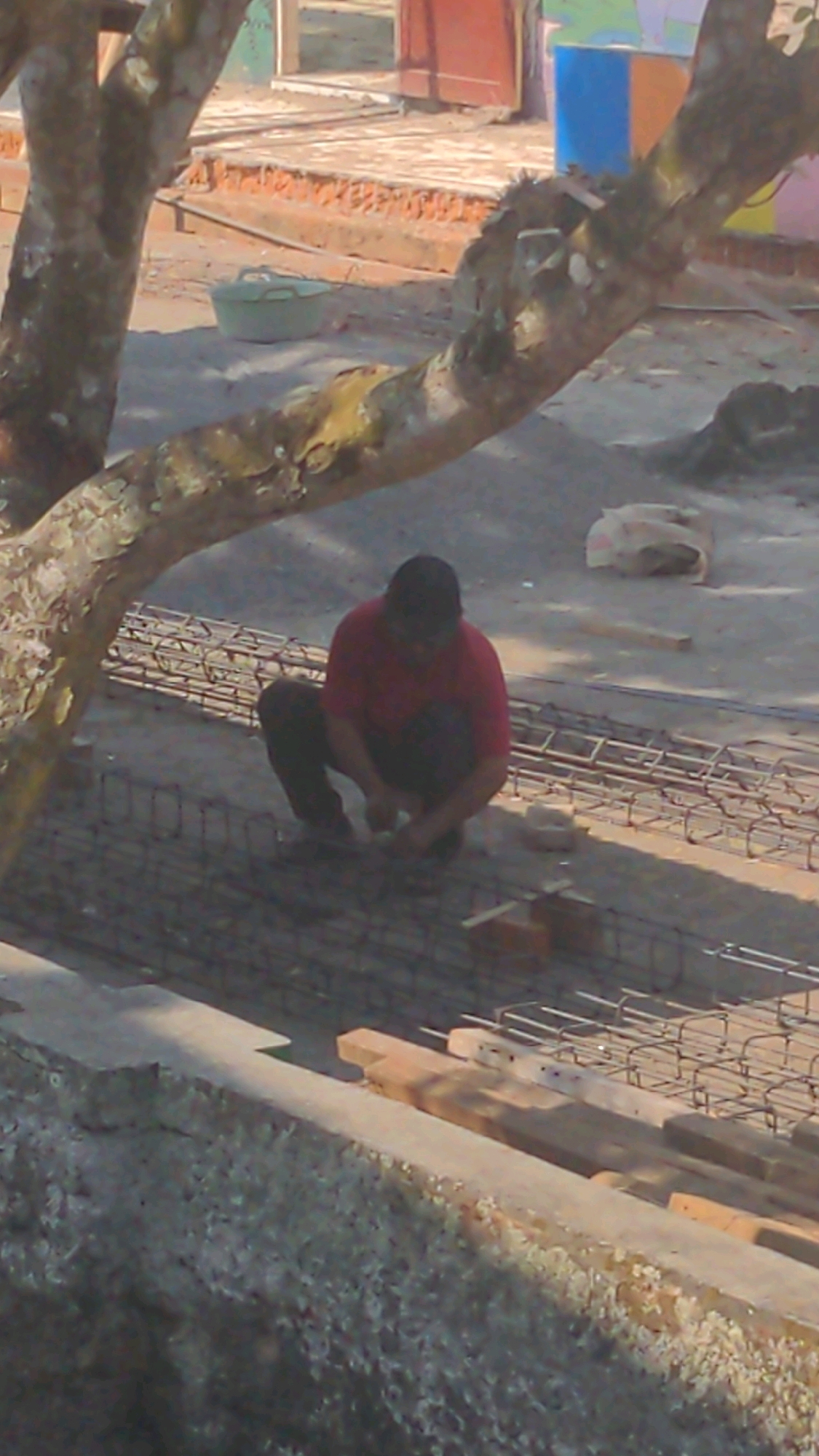 A person wearing a red shirt and dark pants is crouched down, working on what appears to be rebar reinforcement. They are on a construction site with metal rebar laid out in rows. In the background, there are some colorful buildings and a tree. The ground is covered with dirt and gravel. There is a light green bucket to the left of the frame. The image is taken from an elevated perspective.