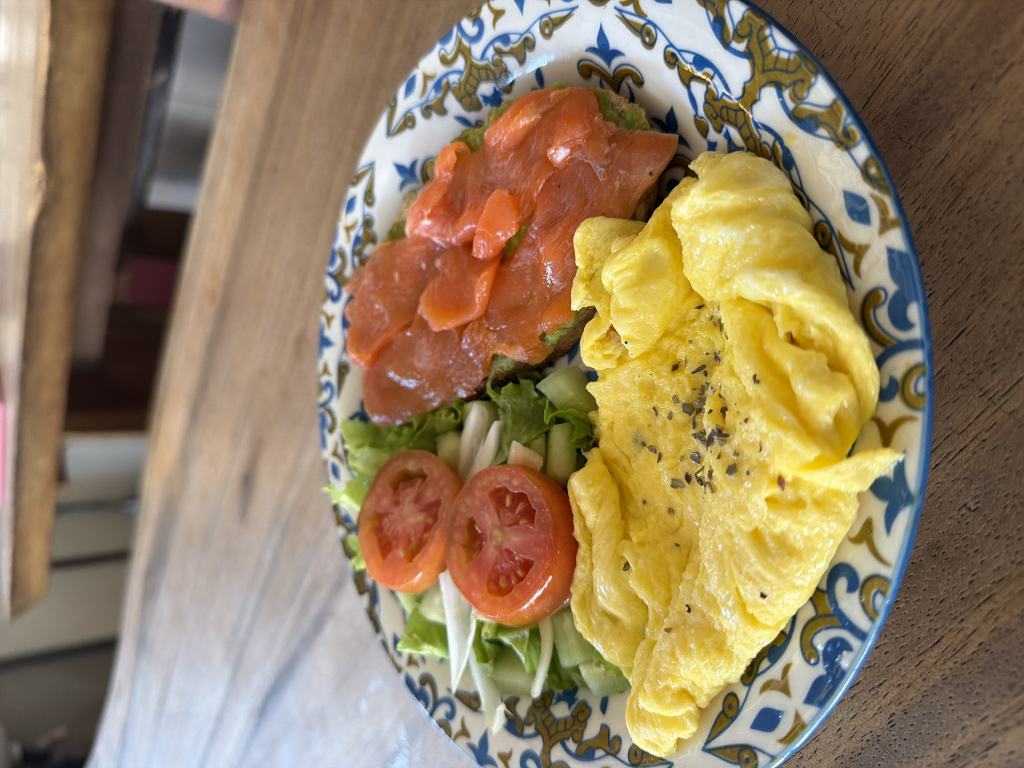 This image presents a close-up, slightly aerial view of a plate of food, presumably a breakfast or brunch, resting on a wooden table. The main subject is a round, white ceramic plate adorned with an ornate blue and golden-yellow pattern around its rim.
The plate holds a visually appealing array of ingredients. On the left side, several bright orange-pink slices of smoked salmon are generously piled atop what appears to be a green base, possibly avocado spread on toast. Below the salmon, a fresh salad garnish consists of vibrant green lettuce, thinly sliced white vegetables (likely onion or cucumber), and two neatly cut, reddish-orange tomato slices. Occupying the right half of the plate is a substantial serving of fluffy, light yellow scrambled eggs, seasoned with visible dark specks, possibly dried herbs or black pepper.
The immediate background is a natural wooden table surface, characterized by its grain and warm brown tones. The background beyond the table is blurred, showing indistinct shapes and varying shades of brown, suggesting an indoor environment with other wooden furniture or structures.
No people are visible in the image, and there is no discernible text. The scene captures a prepared meal, implying the activity of dining. While the location context is Kuta, Indonesia, this detail is not visually evident from the image itself. The meal's composition points towards a daytime setting, likely morning or early afternoon.