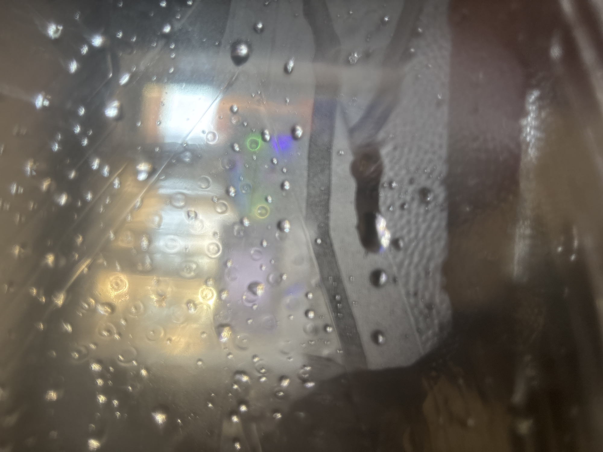 An overhead shot focuses on a dark, wet surface, likely plastic, with numerous small water droplets scattered across it. The droplets create distorted reflections of a brightly lit, colorful background, suggesting the presence of illuminated objects or screens. In the center of the frame, a dark, elongated object, possibly a piece of lint or debris, is visible. The overall impression is that of a close-up view through a wet, transparent material.