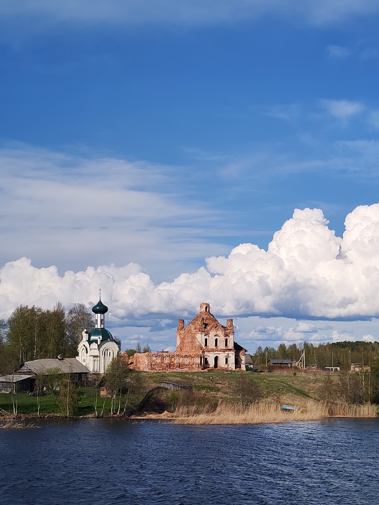 The image depicts a scenic view of Анхимово, Russia, featuring a body of water in the foreground and a clear sky with scattered clouds above. Prominently positioned on the shoreline is a ruined brick building next to a well-maintained white church with a green dome. A small, wooden house is adjacent to the church. A small boat lies near the shoreline on the right side of the image, amidst dried reeds. The environment is a rural landscape with trees dotting the horizon and lush greenery.