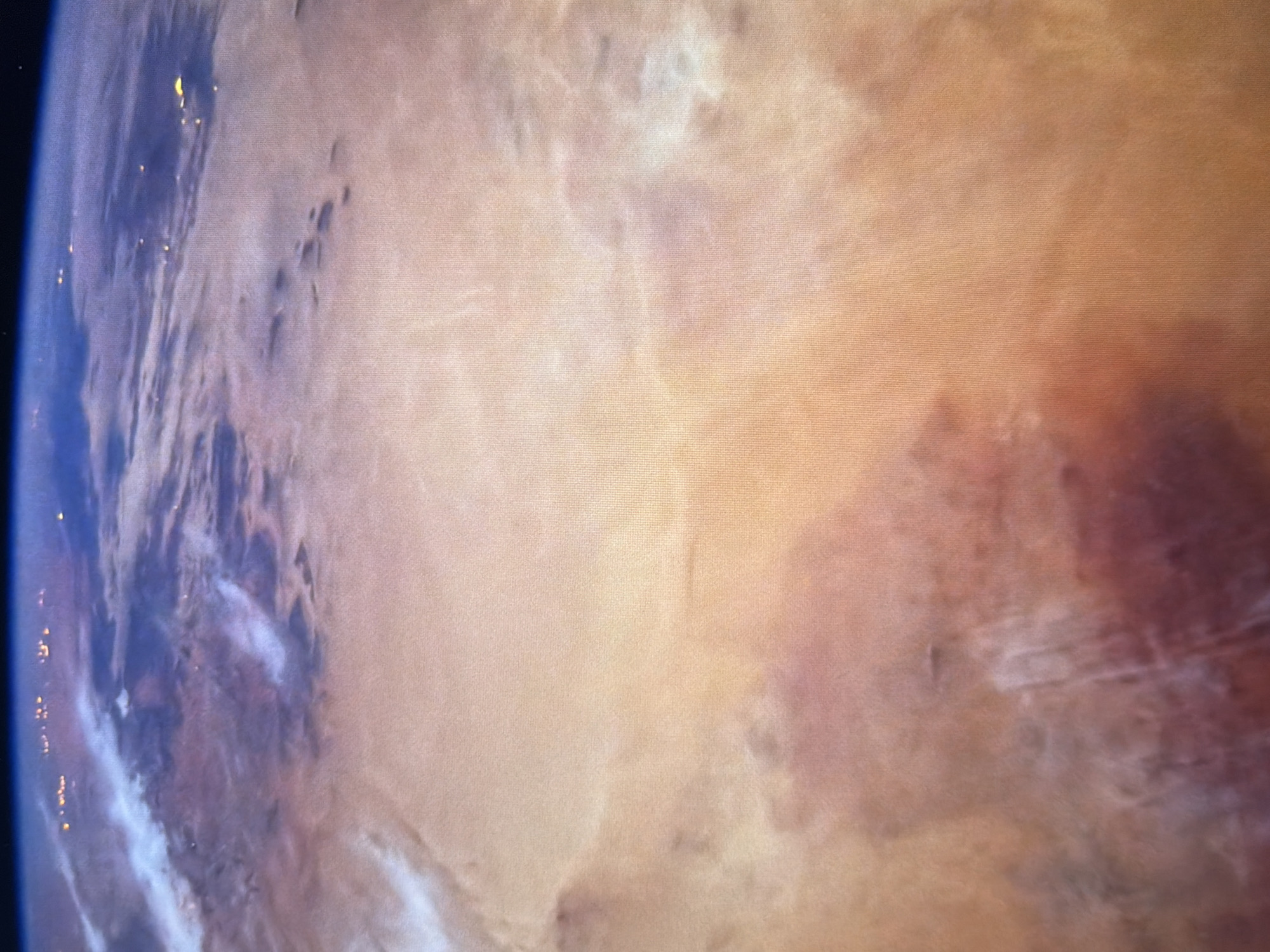 The image is a satellite view of Earth. The upper portion of the image shows a curved horizon with scattered lights, possibly indicating a city at night. Below the horizon, clouds are visible, casting shadows on the land below. The land surface appears to be a desert or arid region, with varying shades of brown, orange, and red. Some areas show lighter patches, which could be sand dunes or geological formations. There are also some darker areas that might represent mountains or dried riverbeds. The overall impression is of a vast, uninhabited landscape seen from a high altitude. The image does not show any specific urban areas that can be identified as Kyiv, Ukraine, nor does it depict any people, objects, or actions.