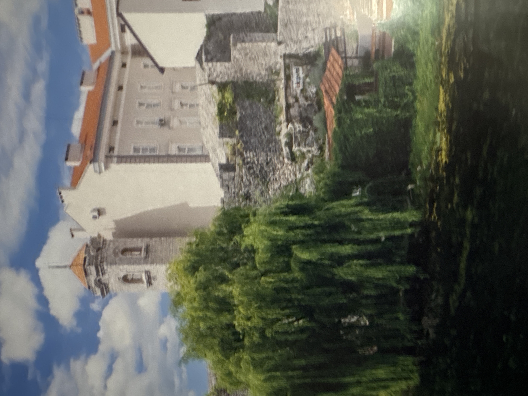 The image displays an architectural scene, likely an old town or fortress, under a bright blue sky with dispersed white clouds. In the left foreground, a large, vibrant green deciduous tree with characteristically weeping branches dominates the lower section. Above and to the right of the tree, a light-colored stone tower features a pointed, reddish-orange tiled roof and multiple arched windows. Adjacent to the tower, a multi-story building, pale beige in color, presents numerous rectangular windows and a red-tiled roof, with visible air conditioning units mounted on its exterior. Below these structures, extensive dry-stacked stone walls and terracing are visible, suggesting fortifications or a hillside development. Further to the right and at a lower elevation, a smaller structure with a reddish-orange tiled roof is almost entirely covered by dense green ivy or vines. No people, vehicles, or specific identifying signage are discernible in the scene. The precise location within the city of Unknown, Unknown, is not identifiable from the image content.