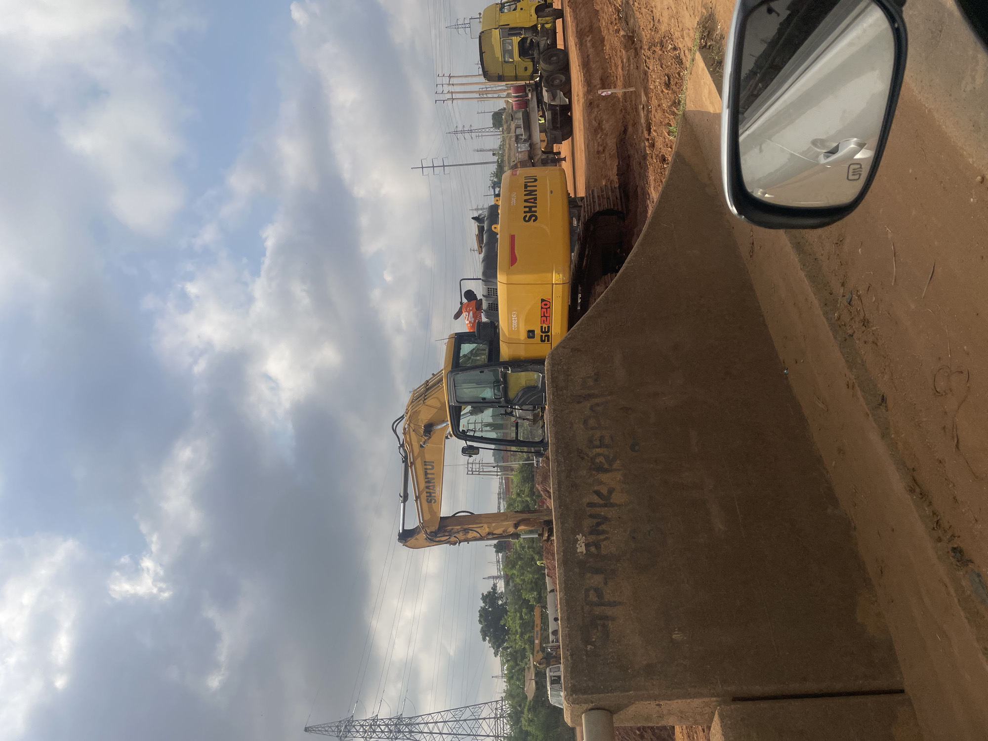 The image shows a construction site. A yellow SHANTUI SE220 excavator is positioned next to a concrete barrier on which "TANK REPAIR" is written. A truck and trailer are parked behind the excavator. Power lines and towers are visible in the background. The image appears to be taken from inside a vehicle, with the side mirror visible. The sky is cloudy. I cannot confirm the location as Abuja, Nigeria.