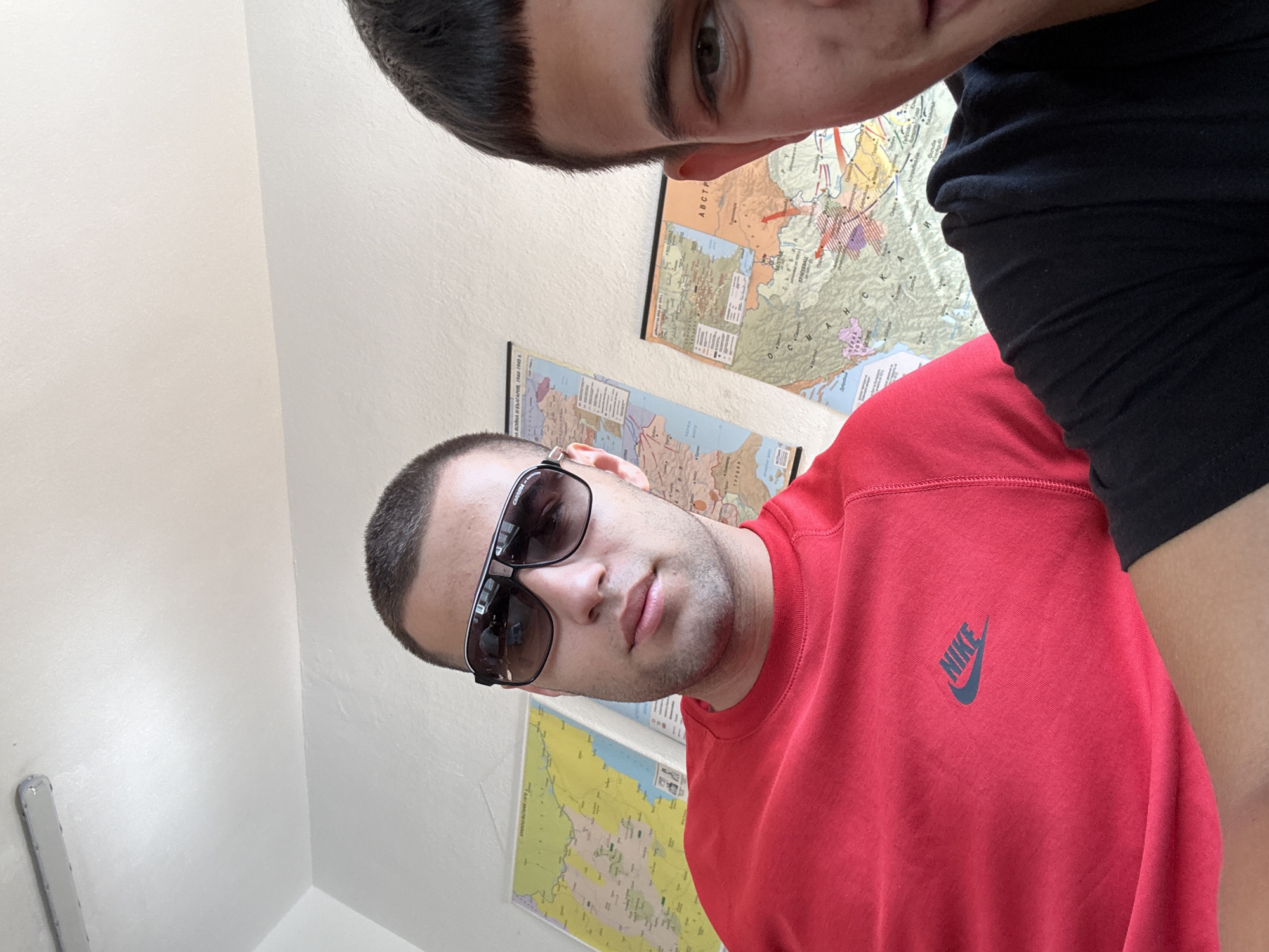 The image shows two young men indoors. The man in the foreground, on the left, is wearing a red Nike sweatshirt and sunglasses. He is looking towards the camera. Behind him, on the wall, are two maps. The man on the right is partially visible on the right side of the frame, wearing a black t-shirt and looking towards the camera. The environment appears to be a room with white walls and a fluorescent light fixture on the ceiling. The maps on the wall are geographical, with one appearing to be a map of the Balkan region, possibly including Bulgaria. Based on the maps and the context provided, the location is the city of Plovdiv, Bulgaria.