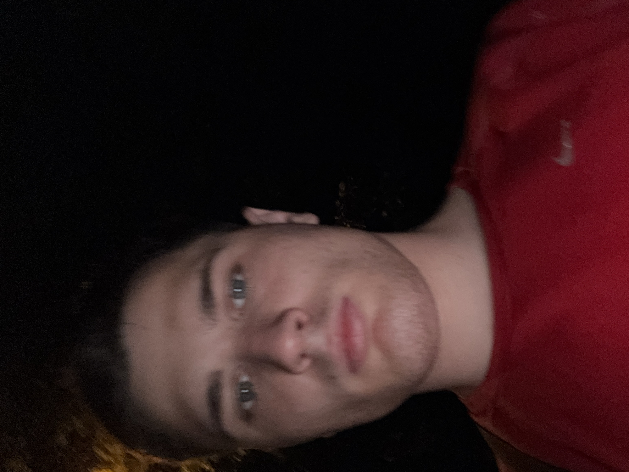 A close-up, night-time, outdoor selfie features a young man with light skin, brown hair, and blue eyes. He is wearing a red t-shirt with a white Nike logo on the left breast. The background is dark, suggesting an urban or semi-urban environment at night. There is a faint yellow-orange light source visible on the left side of the image, possibly from streetlights or foliage. The man's expression is neutral. The image was taken in Stara Zagora, Bulgaria.
