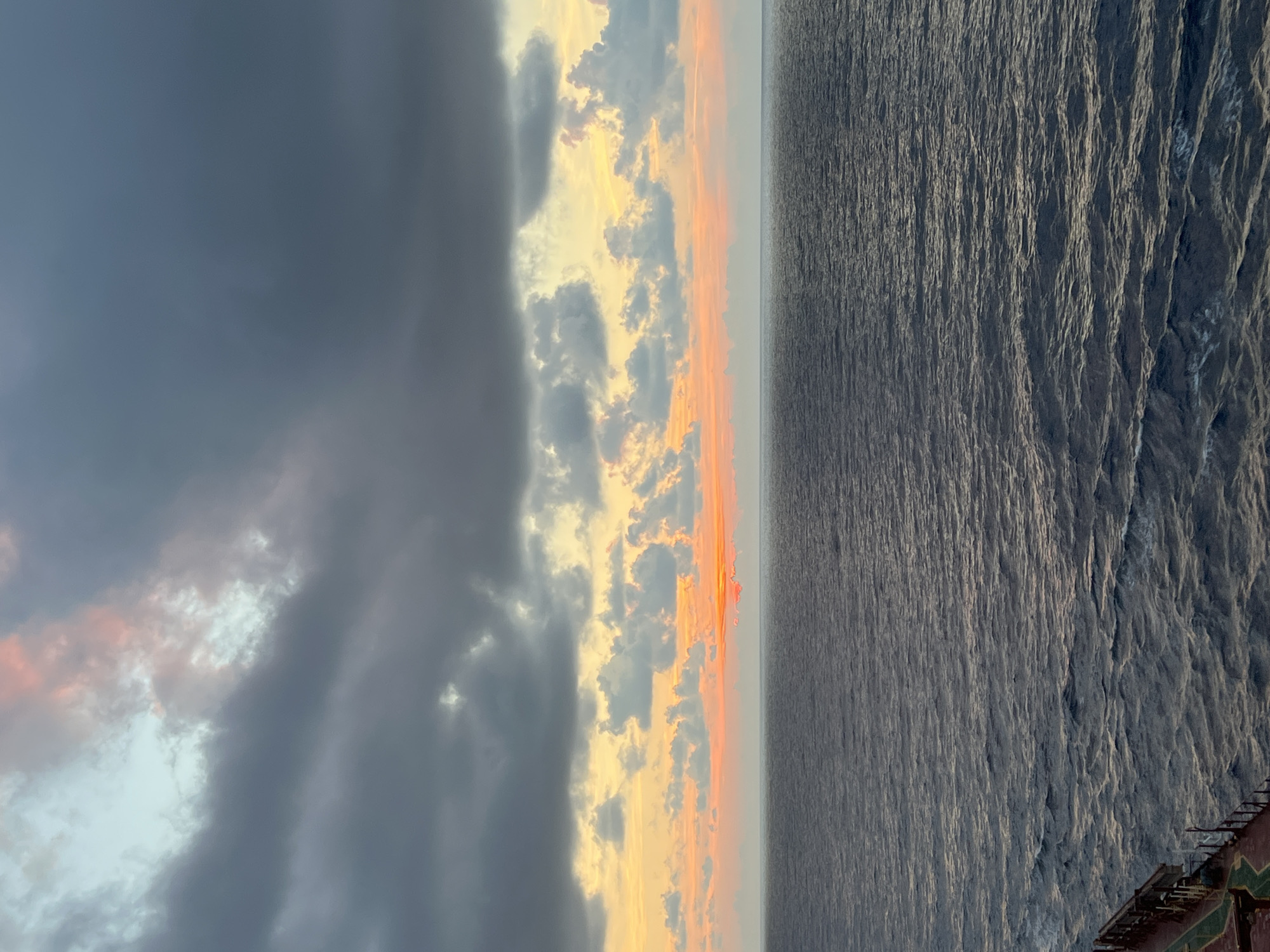 The image depicts a sunset over a vast ocean. The sky is filled with dramatic clouds, ranging from dark, stormy gray to lighter shades tinged with pink and orange. A band of warm color, likely from the setting sun, is visible along the horizon, partially obscured by clouds. The ocean below is a deep blue-gray, with gentle waves creating a textured surface. In the bottom left corner, a portion of a ship's railing and deck is visible, suggesting the photo was taken from a vessel at sea. The overall impression is one of natural beauty and the immense scale of the ocean and sky.