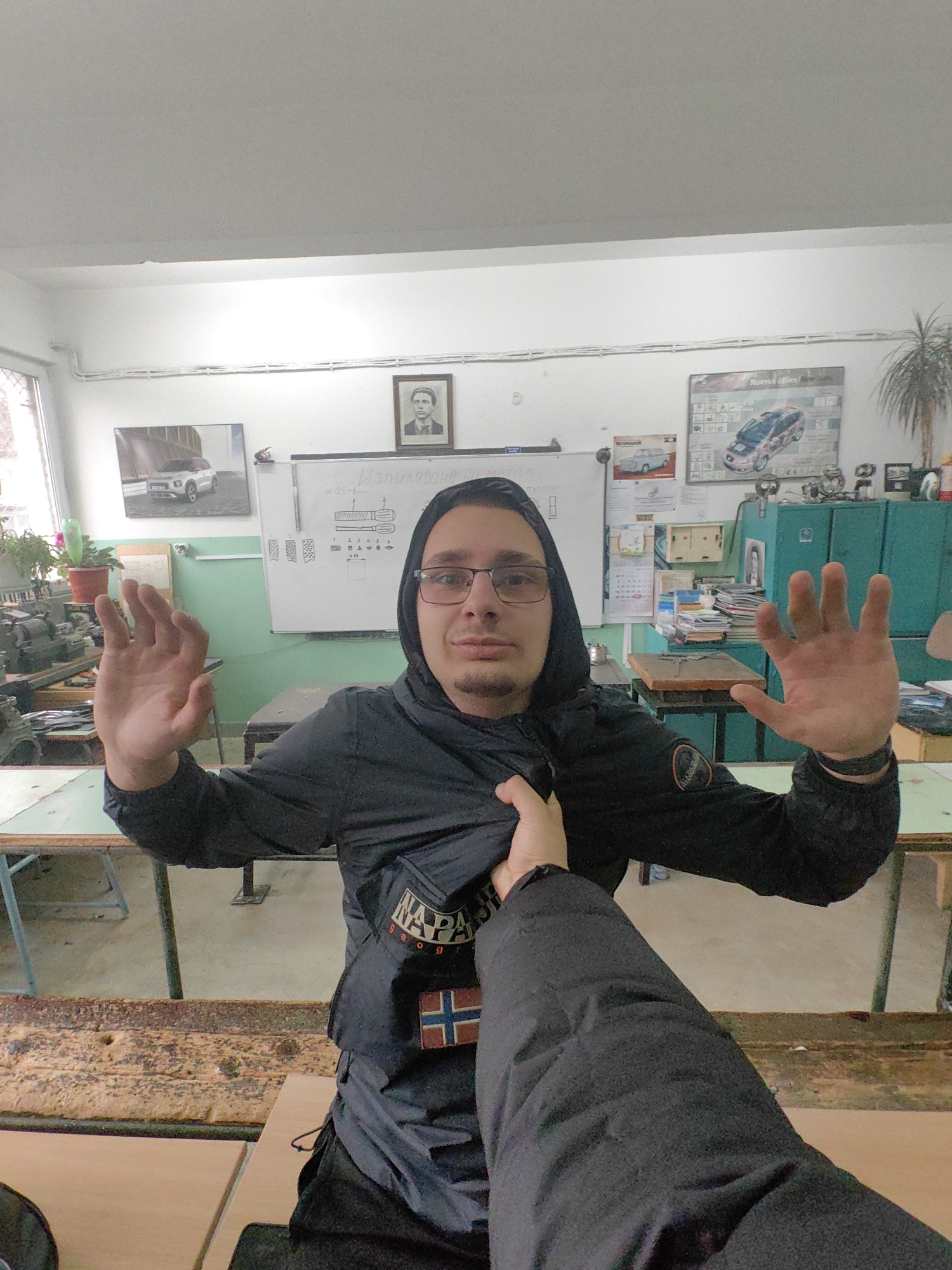 A medium, indoor shot depicts a man in a classroom or shop-like environment, facing the camera with his hands raised. He wears glasses and a black hooded jacket with a "Napapijri" patch and Norwegian flag emblem on the front. A hand is reaching for his jacket. The classroom contains wooden desks, a whiteboard with diagrams, and posters of cars, including a Citroen C4. A framed portrait hangs above the whiteboard. The environment appears to be in Sofia, Bulgaria based on the presence of Bulgarian text on the whiteboard and overall classroom style.