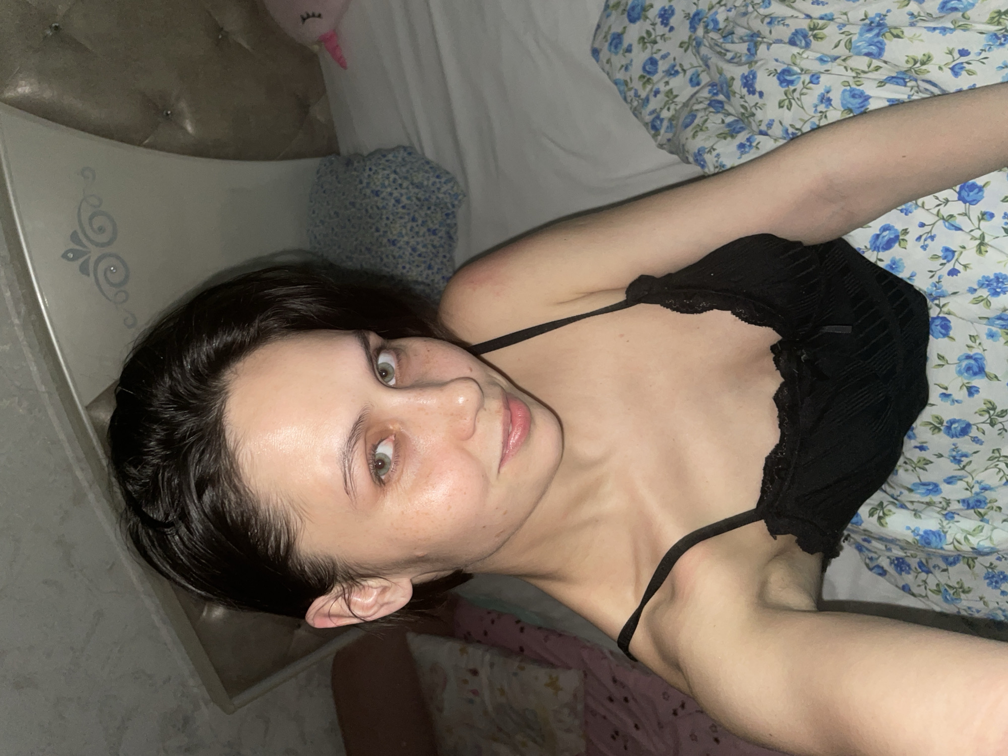 A young woman is captured in a close-up portrait, lying on a bed in what appears to be a bedroom in Izhevsk, Russia. She is positioned on her side, looking directly into the camera with a neutral, engaging expression. Her dark hair, which appears somewhat damp, frames her face. Her greenish-grey eyes are open, and her fair skin shows a scattering of freckles across her nose and cheeks. She is wearing a black spaghetti strap top featuring lace details.
The setting suggests an indoor environment, likely a bedroom, with subdued lighting indicative of evening or nighttime. The bed is covered with a blue floral patterned duvet or sheet featuring blue roses, and a matching patterned pillow is visible behind her head, alongside a plain white sheet. In the background, an ornate, light-colored headboard with tufted detailing and a side panel featuring a silver swirl design is visible. A small pink unicorn plush toy is partially seen near the top of the bed. No text is visible in the image.