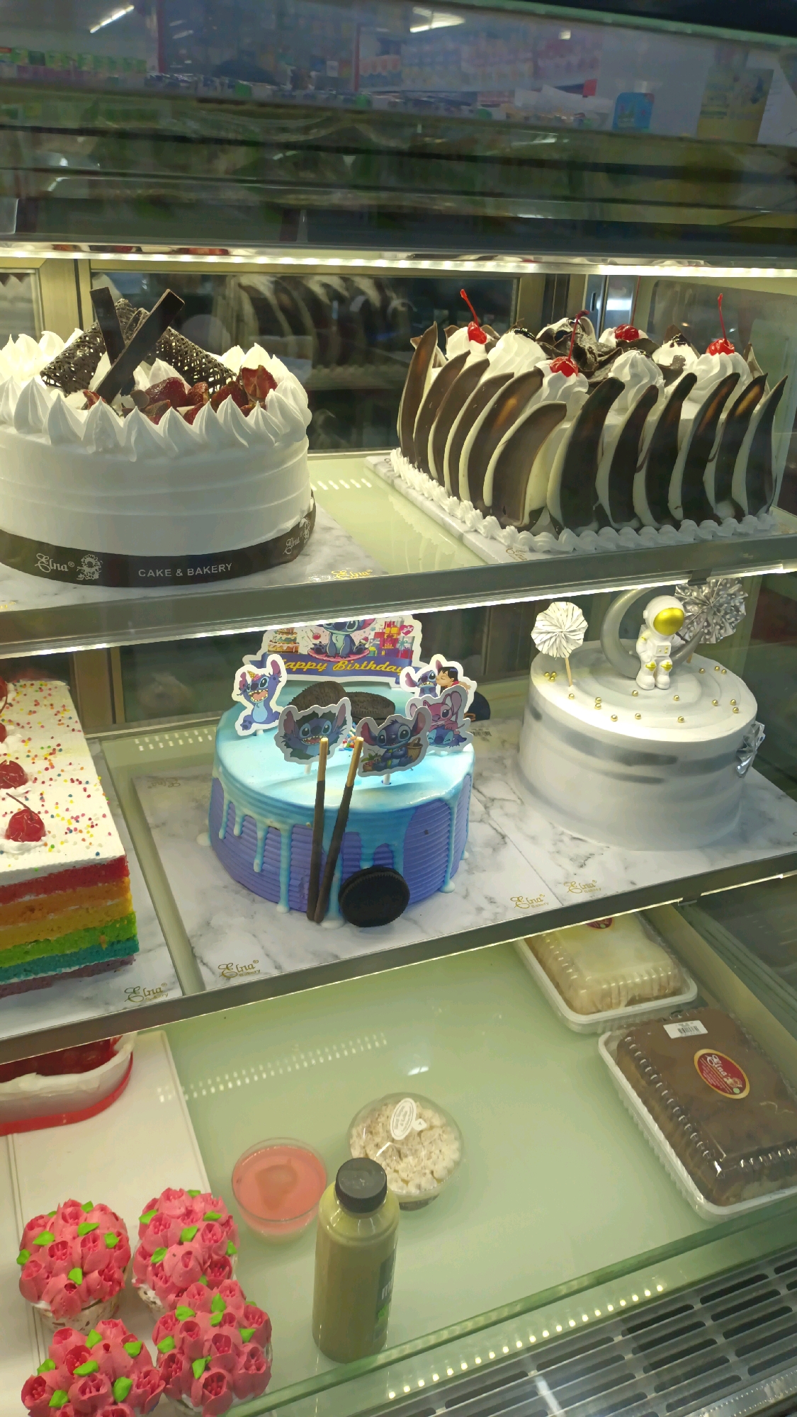 The image displays the interior of a bakery, likely in Padang Panjang, Indonesia, with a selection of cakes and other desserts showcased in a refrigerated display case.
In the top tier of the display case, a large white cake is decorated with white frosting swirls, chocolate pieces, and strawberries. To its right, another cake features a similar white frosting base adorned with chocolate leaf-shaped decorations and topped with cherries.
The middle tier showcases more cakes. On the left, a slice of a rainbow layered cake with white frosting and cherries is visible. Next to it is a blue cake with dripping frosting, decorated with cartoon characters, two brown sticks, and an Oreo cookie. To the right of the blue cake is a grey and white cake featuring an astronaut figurine on top, a crescent moon, and silver starburst decorations.
The bottom tier of the display case contains several items. On the left are several pink cupcakes with green frosting accents, arranged in a cluster. Adjacent to them are a small bowl of pink liquid, a clear plastic container with a white crumbly dessert, and a green bottle with a black cap, possibly a beverage. Further to the right, there are two rectangular cakes in plastic packaging, one lighter in color and the other a dark brown cake.
The overall environment suggests a clean and well-lit retail space, typical of a bakery selling freshly made goods. The reflections in the glass display case indicate the presence of shelves and items in the background, though they are out of focus.