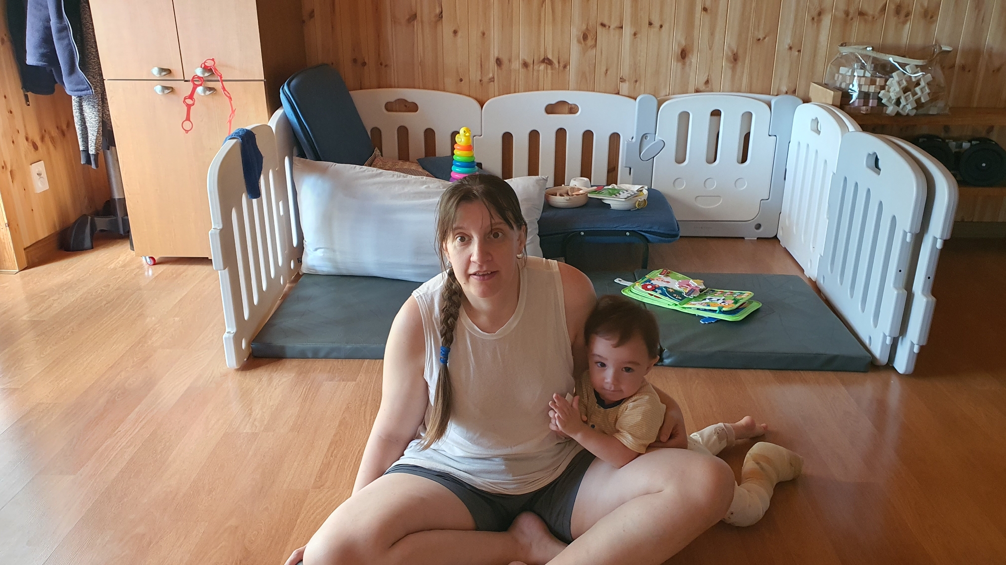 The image depicts a woman and a baby sitting on a wooden floor within an indoor space, likely a residence in Dunnae-myeon, South Korea. The woman is fair-skinned and wearing a white tank top and dark shorts; her hair is in a braid. The baby, who has darker hair and skin, is wearing a striped yellow shirt and light-colored leggings.
Behind the woman and baby is a play area bordered by white plastic play yard panels. Within the play area are several cushions, a pillow, and various baby toys, including a stacking toy and a soft book.
To the left of the play area is a light-colored wooden cabinet with clothes hanging on its left side. The floor is light-colored wood. The background wall appears to be paneled with light-colored wood planks. The lighting is bright and appears to be natural.