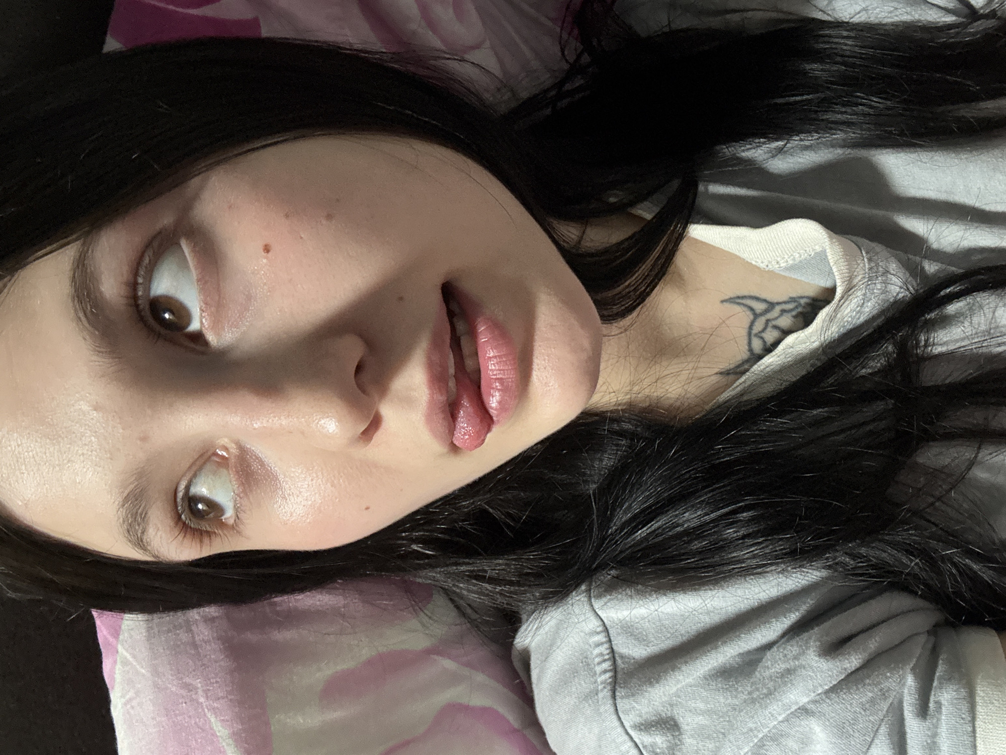 A woman with long, dark hair and fair skin is lying down. Her head is on a pink and white patterned surface that appears to be a pillow or bedding. She is wearing a light gray top. She has brown eyes and a tattoo visible on her upper chest near her neck. Her lips are slightly parted. The location of Фоки, Russia is not apparent in the image.