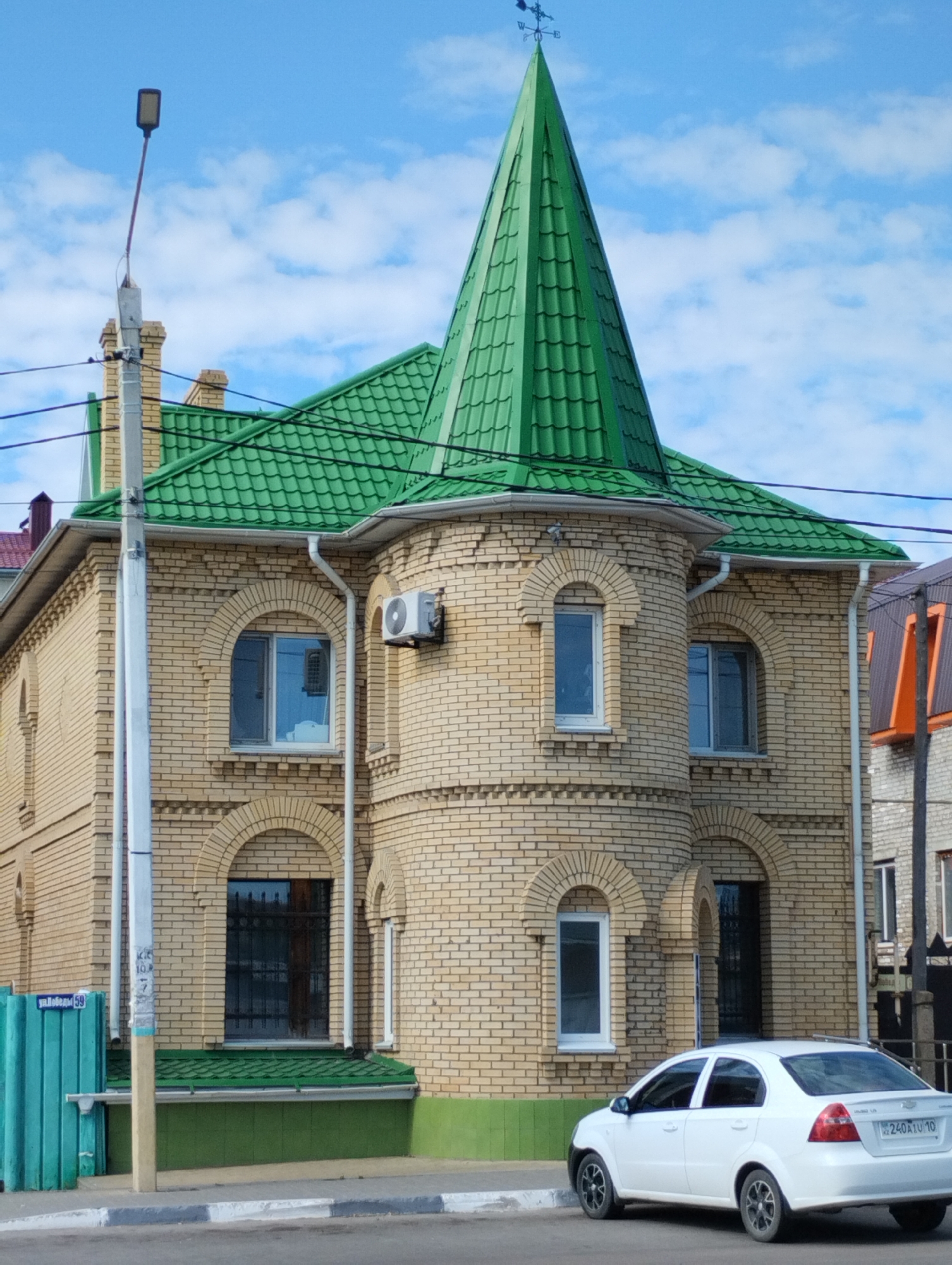 The image shows a two-story building with a prominent green, conical roof and a weather vane on top. The building is constructed of light-colored brick and features arched windows and an outdoor air conditioning unit. A white sedan is parked on the street in front of the building. The street is paved and bordered by a curb. A turquoise fence and a lamppost are visible to the left of the building. The sky is blue with scattered white clouds. The license plate of the car is visible, reading "240A1U 10". The image is likely taken in Kostanay, Kazakhstan, given the context.