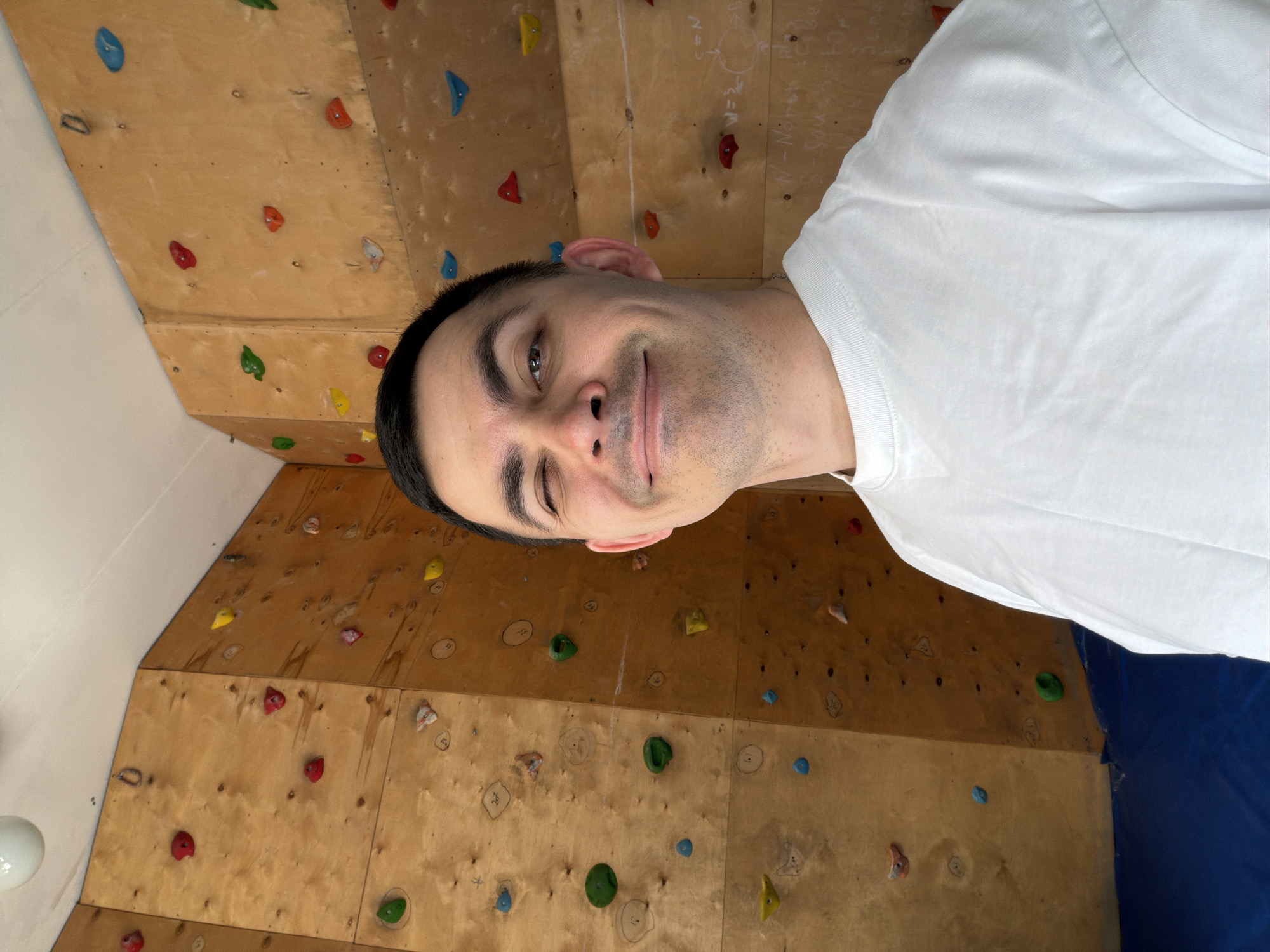 A man is indoors, standing in front of a climbing wall that is constructed of plywood. The wall has multiple climbing holds of varying colors and shapes attached to it. The man is wearing a white t-shirt. His hair is short and dark. He is winking and smiling at the camera. There are some writings in white chalk on the plywood wall. A blue crash pad is positioned below the wall. The location could potentially be in Izhevsk, Russia, but this cannot be definitively confirmed from the image alone.