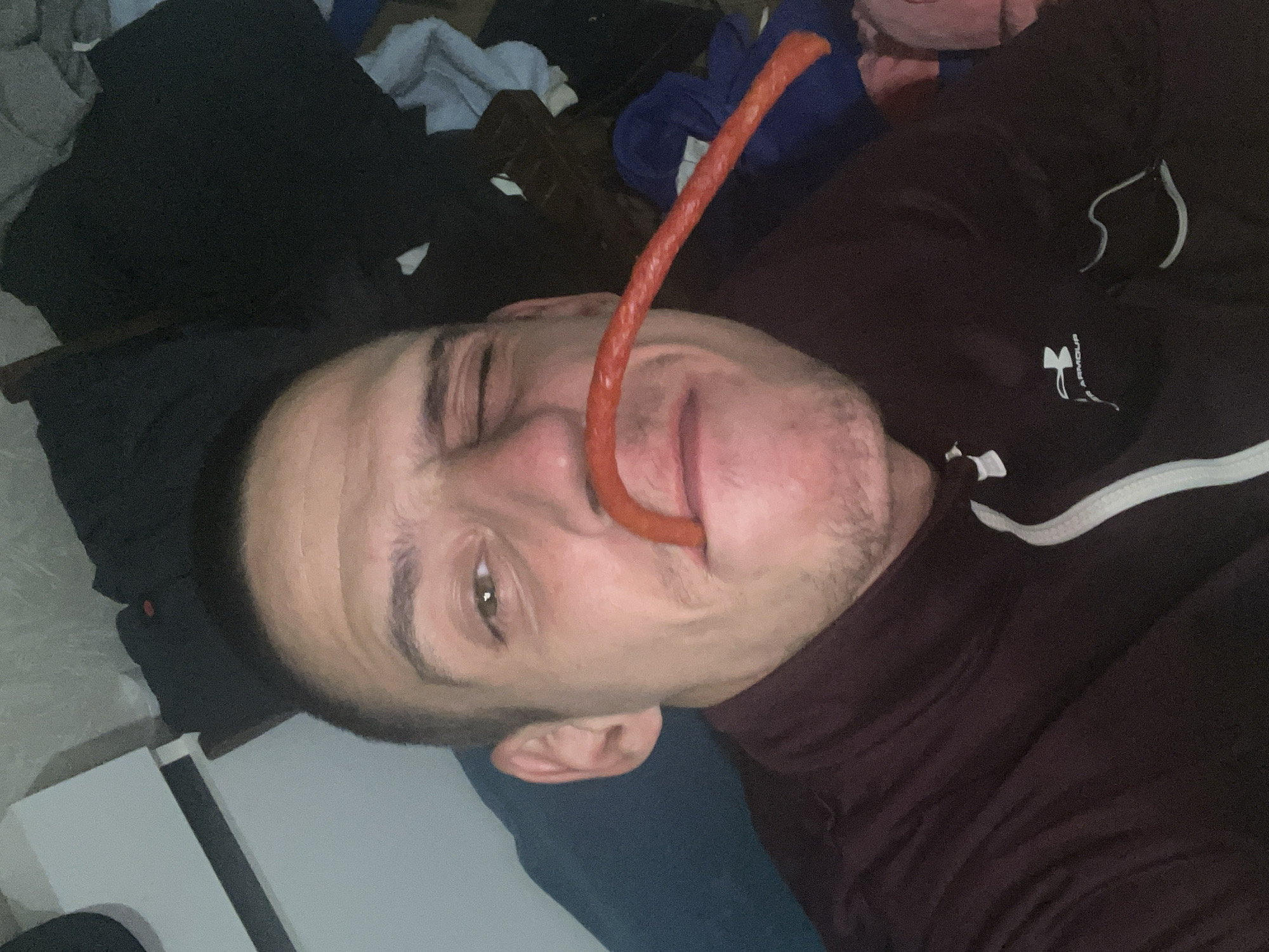 A man is lying down and appears to have a large sausage extending from his nose to his mouth. He is wearing a dark maroon turtleneck sweater with white trim on the collar. The background includes what looks like clothing and other items piled around him. There is no indication in the image that provides any indication of the city of Sofia, Bulgaria.