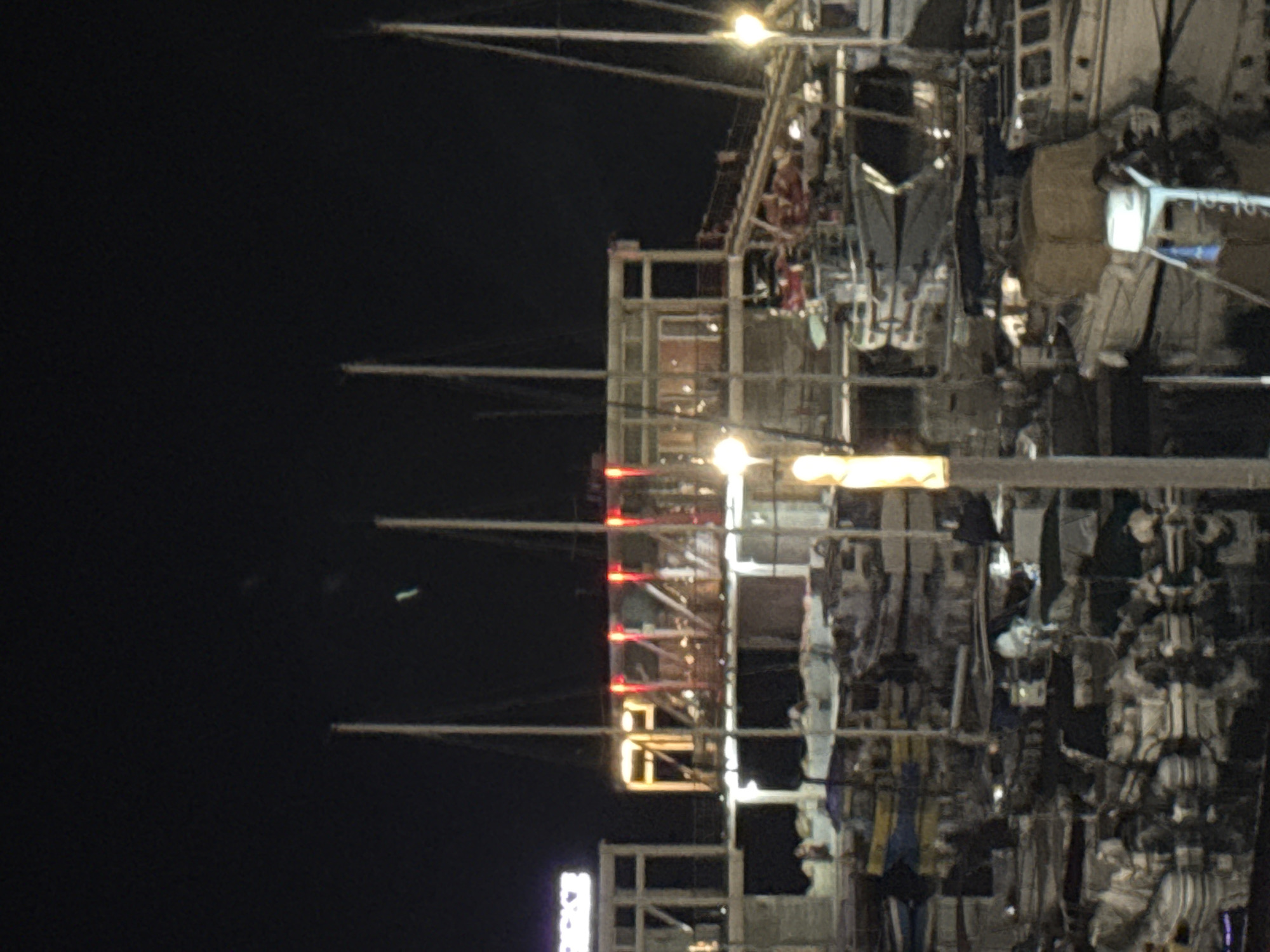 The image depicts a nighttime scene at a marina in Limassol, Cyprus, likely near the old port area or a prominent waterfront development. The foreground is dominated by a reflection in the water, showing numerous boats of various sizes docked and moored. These boats appear to be a mix of pleasure craft and smaller fishing vessels, with some covered by tarps.
The middle ground features a multi-story building, possibly a restaurant or establishment, illuminated with red and white lights. The structure has large windows and balconies, and a ramp or walkway leads up to its entrance. Several masts of sailboats rise above the building.
In the background, the sky is dark and clear. The overall impression is one of a lively, illuminated waterfront at night, with boats reflecting in the still water. The presence of lights and the architectural style suggest a modern urban setting within Limassol.