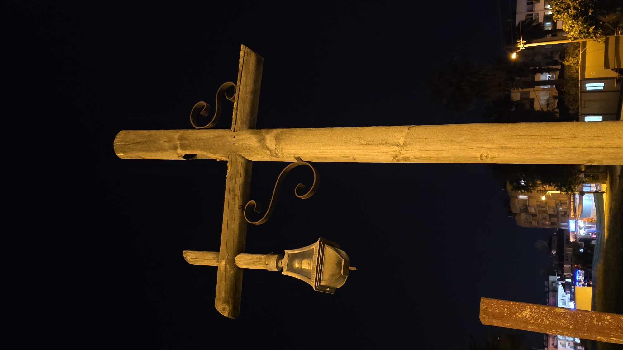 A nighttime view features a wooden signpost with a lamp, likely in Fethiye, Turkey. The signpost is constructed of rough-hewn timbers arranged in a cross shape, with decorative ironwork scrolls. A vintage-style lamp fixture extends from the lower portion of the post. The scene is illuminated primarily by the lamp and background city lights. In the background, buildings and streetlights are visible, indicating an urban setting. The sky is dark, suggesting it is late at night.