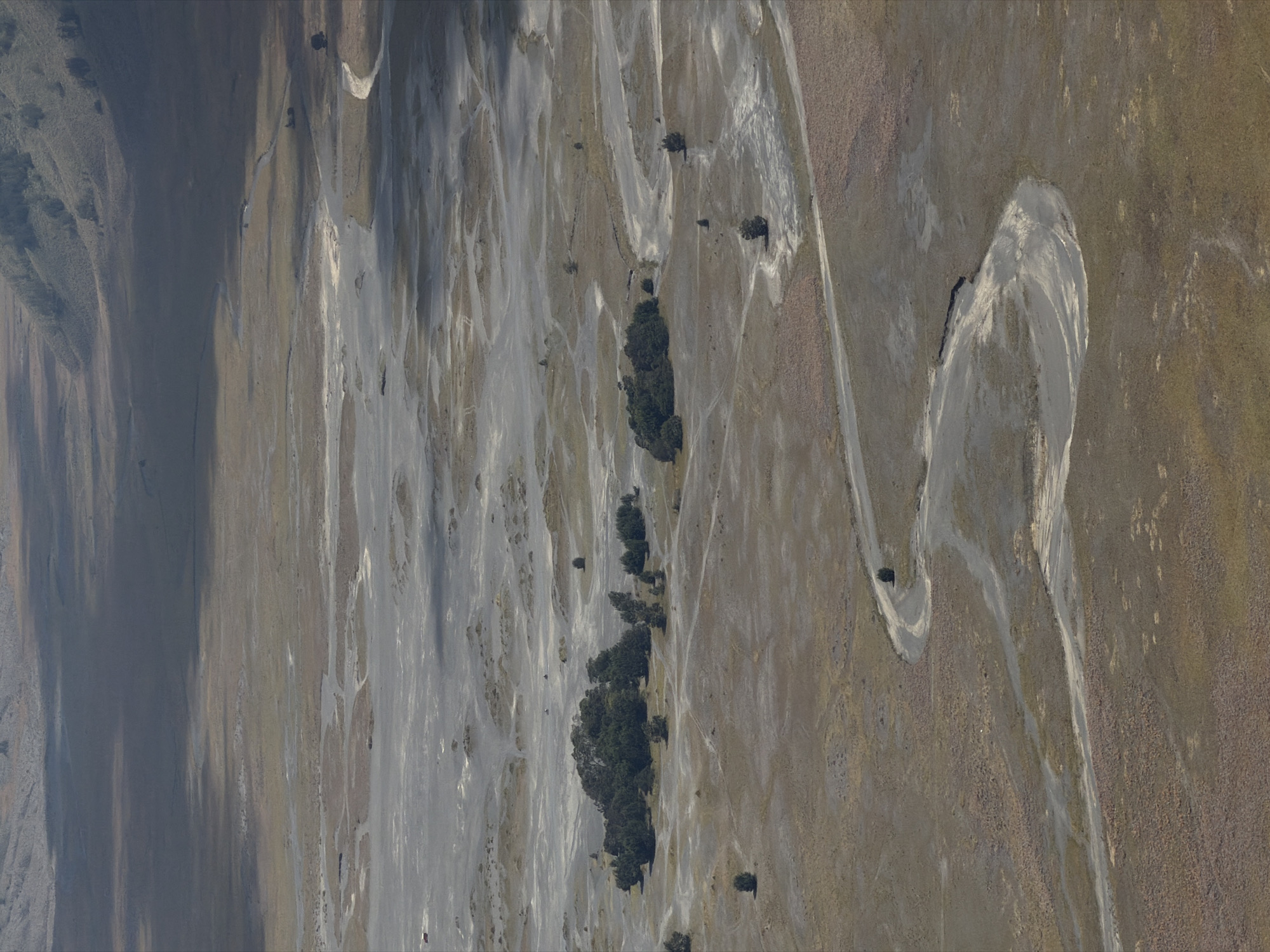 This aerial photograph depicts the vast, arid landscape surrounding Cemorolawang, Indonesia, a region known for its volcanic terrain and dramatic natural beauty. The scene is dominated by a winding, dusty riverbed that meanders through a desolate expanse of dry earth, interspersed with patches of scrubby vegetation. The riverbed itself appears to be a mixture of light gray sand and darker, compacted soil, with evidence of past water flow etched into its surface.
Scattered across the landscape are various geological formations, including low-lying mounds and depressions that suggest erosion and the passage of time. In the mid-ground, a dense cluster of dark green trees stands out, forming a striking contrast against the muted tones of the surrounding environment. These trees, likely a resilient species adapted to the harsh conditions, create a small oasis of life in the otherwise barren terrain.
The sky above is partially obscured by shadows cast by unseen elements, possibly clouds or distant mountains. These shadows create a dynamic play of light and dark across the landscape, accentuating the contours and textures of the terrain. The overall impression is one of a remote and rugged natural environment, shaped by the forces of geology and the elements. There are no visible people or man-made structures, reinforcing the sense of untouched wilderness.