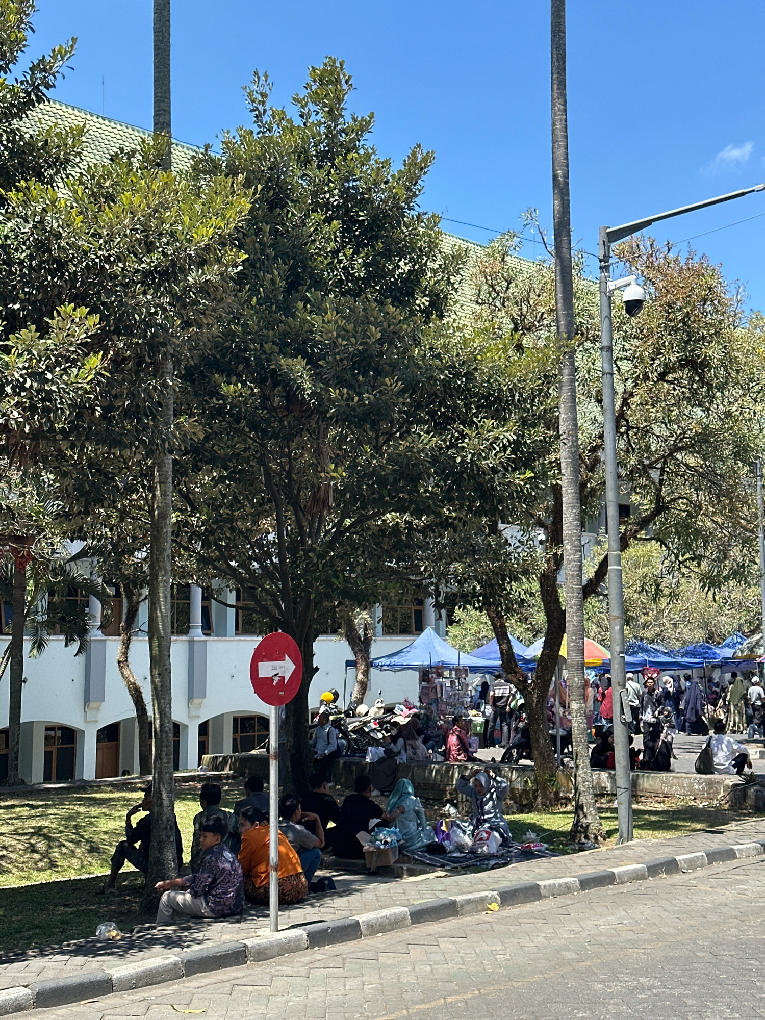 The image depicts an outdoor market scene in Malang, Indonesia, likely on Jalan Semeru, characterized by the presence of stalls and people. A light blue building with white columns and arched windows is visible in the background, suggesting a public or institutional structure. Several people are seated on the ground under the shade of trees, some engaged in conversation or resting, while others appear to be vendors or shoppers at the market. Numerous tents and awnings of various colors, including blue and red, are set up, displaying goods for sale. A traffic sign with a white arrow pointing to the right on a red circular background is prominently placed near the foreground. A CCTV camera is mounted on a lamppost. The ground is paved with cobblestones, and a sidewalk runs along the edge of the street. Several trees with lush green foliage are scattered throughout the scene, providing shade and a natural element to the urban environment. The sky is clear and blue, indicating a sunny day. Motorcycles are parked behind some of the stalls.
