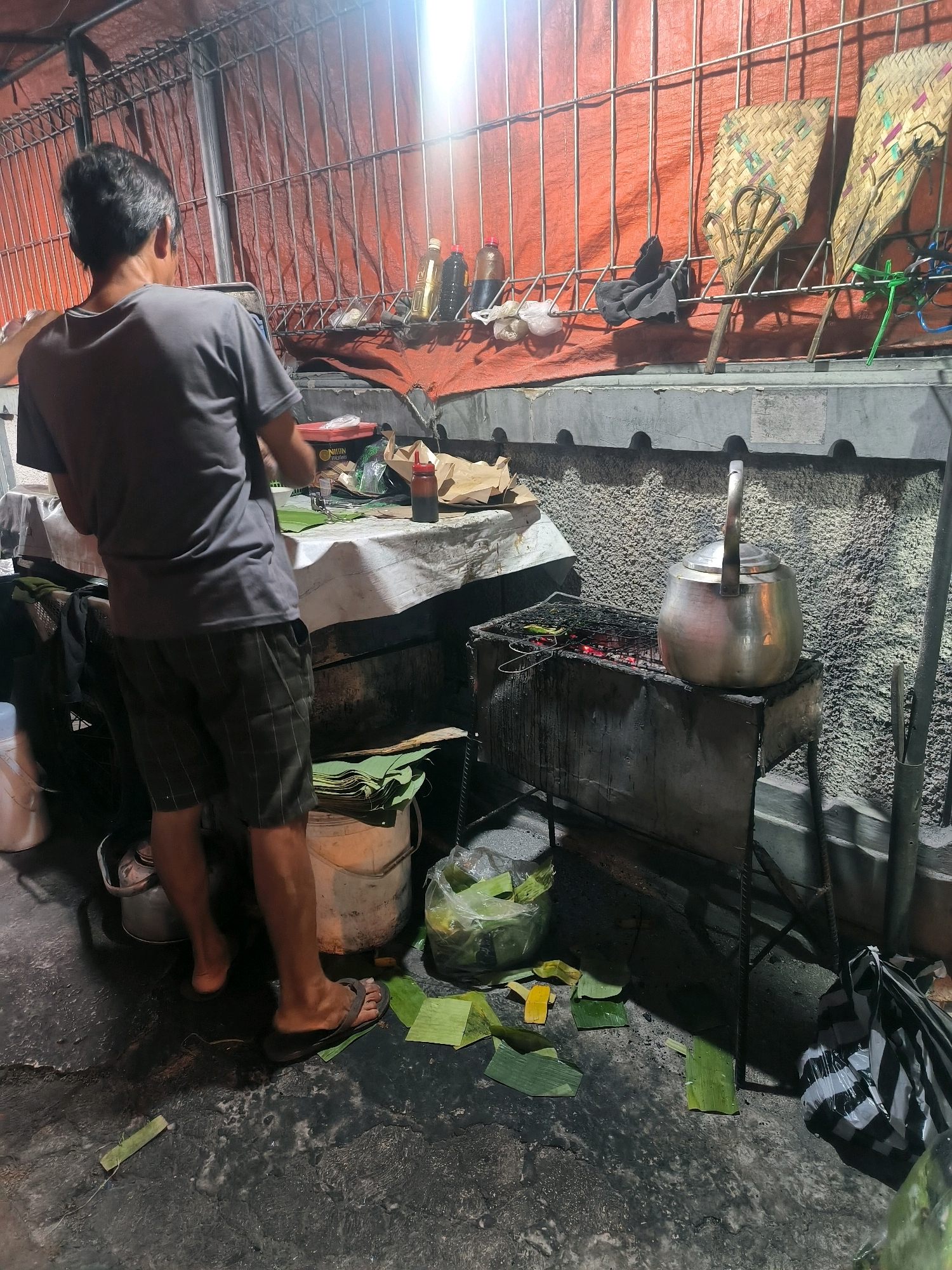 A street food vendor, an adult male with short, dark hair and a grey t-shirt, stands with his back to the camera in Salatiga, Indonesia. He is preparing food on a table covered with a white cloth. Various items are visible on the table, including food preparation materials, bottles of condiments, and bags. In front of the table, a grill with glowing coals is set up, topped with a metal kettle. The surrounding area is paved and appears to be an outdoor market or street stall. Banana leaves are scattered on the ground, some used for food preparation and others fallen. A large plastic bag with items inside and a striped bag are also present on the ground. The background features a metal grate and a draped orange tarp. The overall scene depicts a night-time operation of a food vendor.