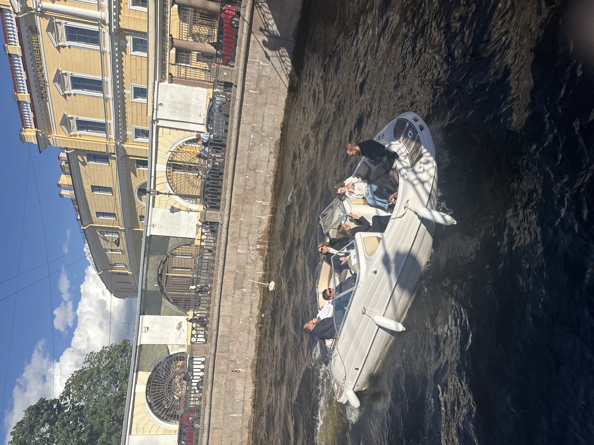 A motorboat with six people on board travels down a canal in Saint Petersburg, Russia. The boat is white with black trim and has several people seated and standing. In the foreground, the water is dark and choppy, with the wake of the boat visible. In the background, a stone embankment lines the canal, topped by a promenade where several pedestrians are visible. Beyond the promenade, a large, ornate yellow building with a classical facade stands under a blue sky with scattered white clouds. The building features multiple windows and decorative architectural elements.
