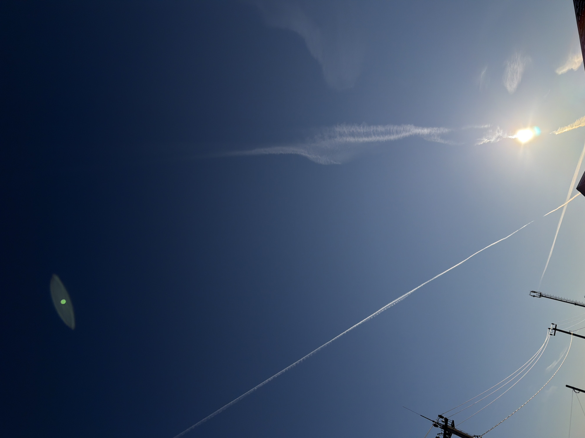 The image shows a bright blue sky with several contrails. A lens flare is visible in the lower left corner. The sun is positioned near the upper right corner, obscured by thin clouds. Power lines and utility poles are visible along the bottom right edge of the frame. The image was likely taken during the day, and shows traces of aircraft vapor in the atmosphere above the location, probably in the city of Chaikovsky, Russia.