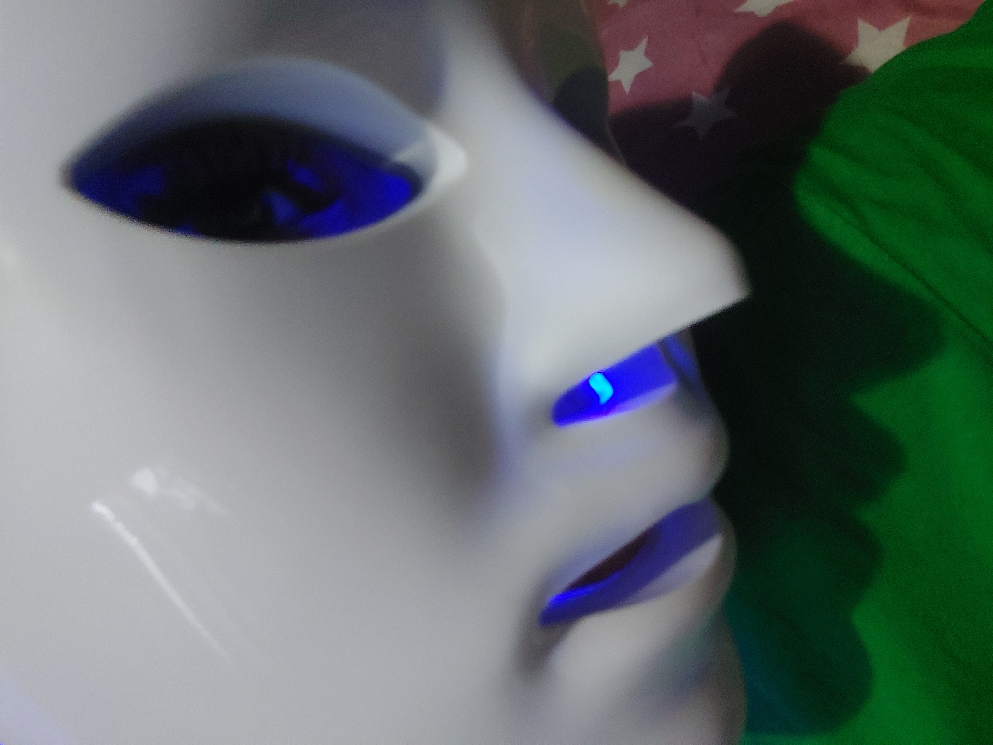 The image shows a close-up, slightly angled view of a white, smooth, mask-like object with sharp, pointed features. There are two prominent areas where blue light emanates, one resembling an eye socket with a dark, reflective surface within, and another smaller point of blue light below and to the left. The mask appears to be resting on a surface covered with a green fabric. In the background, a pink fabric with white stars is partially visible. The lighting suggests an indoor setting, possibly artificial, given the intense blue glow from the mask. The image is slightly out of focus, particularly around the edges and the central portion of the mask.