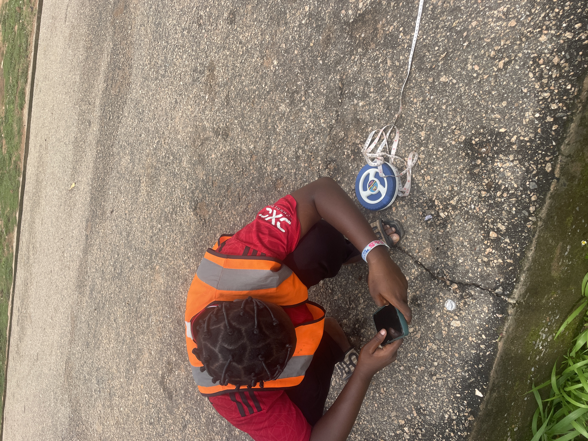 In Lokoja, Nigeria, a person wearing a red shirt and an orange reflective vest is crouched on a paved surface, examining a smartphone. Beside them lies a blue and white measuring tape, partially unspooled across the ground. The person's hair is styled in numerous small braids. The environment appears to be outdoors, with the paved area showing signs of wear and cracks, and a strip of green grass bordering the lower edge of the frame.