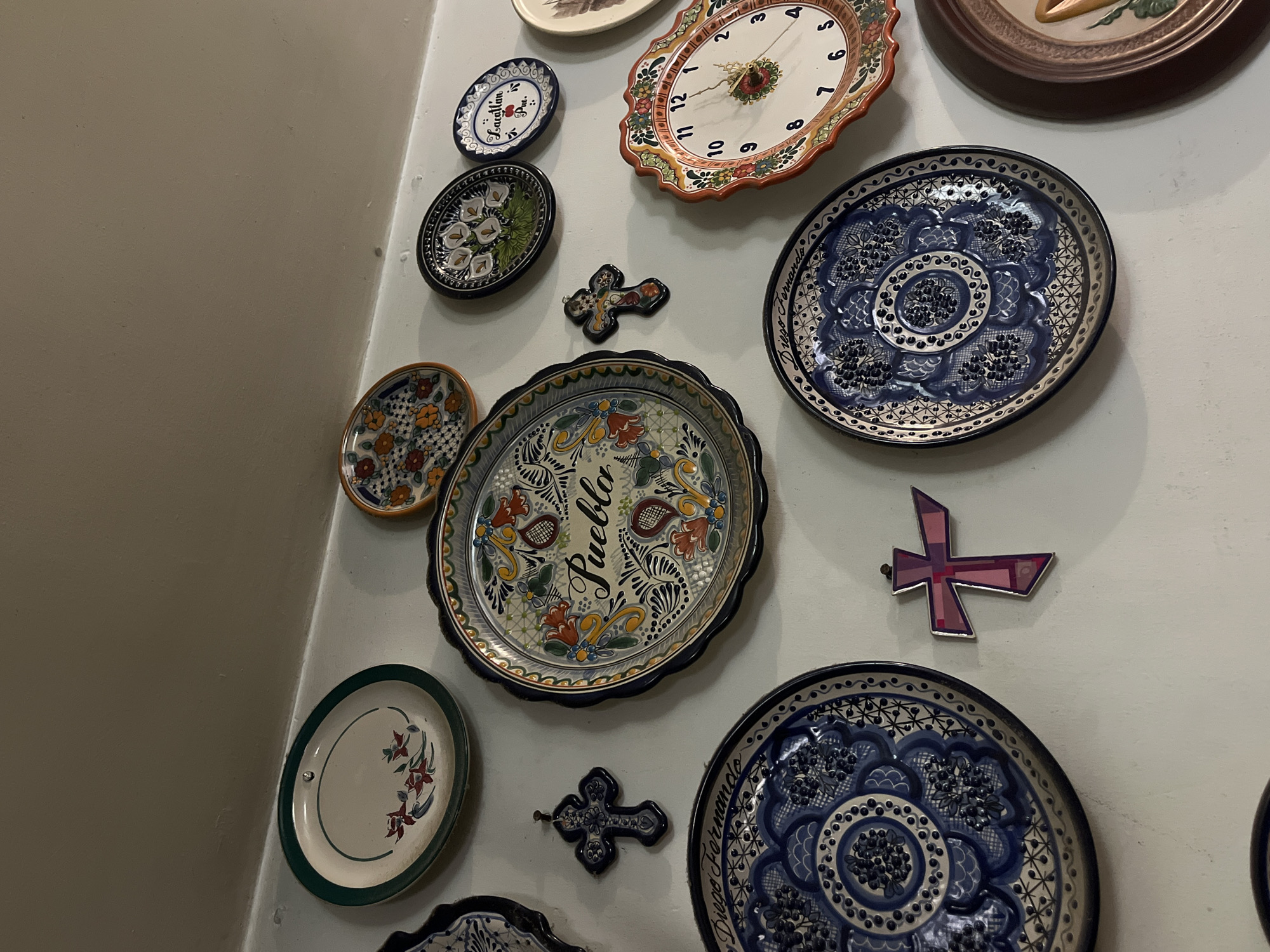 A collection of decorative plates and crosses are mounted on a white wall. The plates vary in size, color, and design, with some featuring floral patterns, geometric designs, and the word "Pueblo." One plate is actually a clock. The crosses are made of different materials, including metal and ceramic. The plates and crosses are arranged in a somewhat haphazard manner, creating a visually interesting display. The location appears to be inside a building, possibly a home or museum.