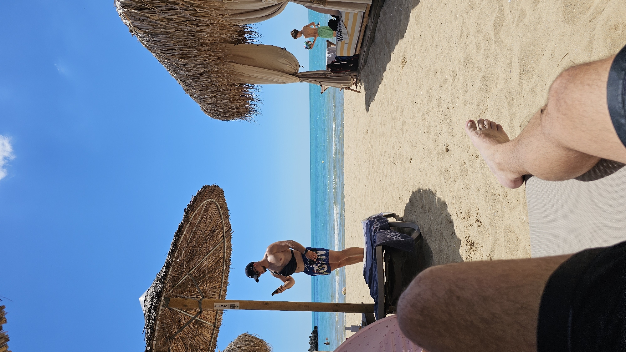A man is standing on a beach in Varna, Bulgaria, holding a smartphone. He is wearing blue and white patterned shorts, a black fanny pack, and a black baseball cap. He appears to be looking at the phone. In the background, there are thatched umbrellas and beach chairs. The sea is visible, with a clear blue sky above. The sun is shining, indicating it is daytime. The sand is light brown. There is a child in the distance to the right of the man, standing near a beach cabana. The image is taken from a low angle, looking up towards the sky and the beach umbrellas. Another person's legs and feet are visible in the foreground, suggesting the photo was taken by someone sitting on a beach chair. A blue towel is draped over a beach chair.