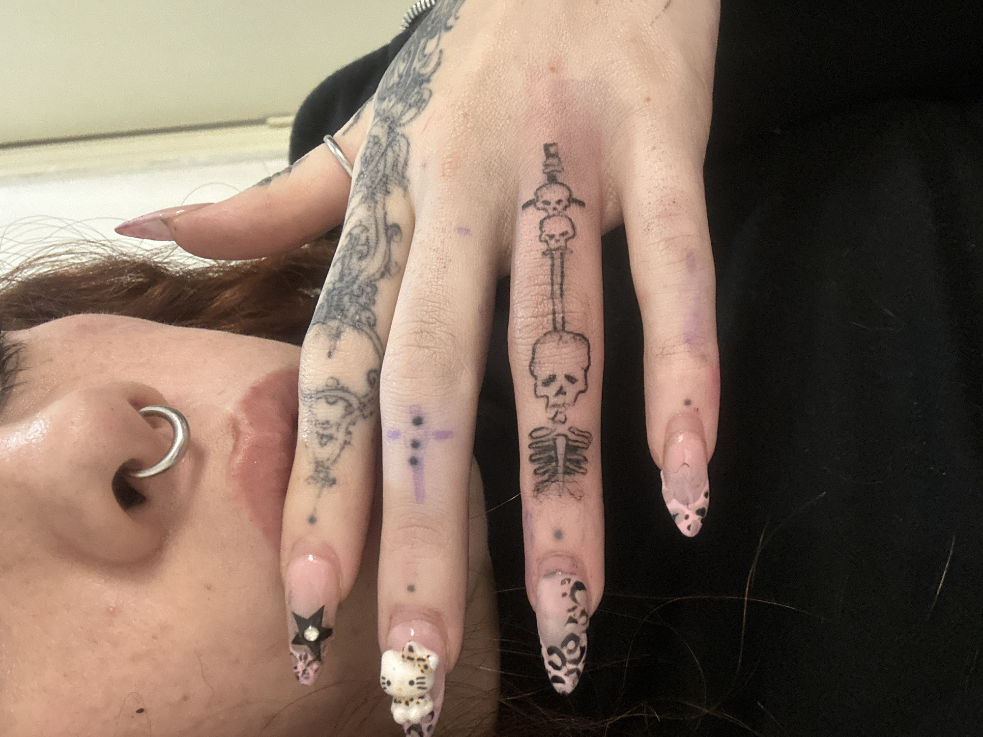 A close-up shot depicts a person's hand and part of their face. The hand features various tattoos on the fingers and back of the hand. The fingers also have manicured nails with different decorative elements, including a star, Hello Kitty, and a leopard print. The face shows a person with a nose ring. The background is blurred, but a light-colored surface can be seen. The location is undetermined as there is no identifiable information to suggest it is in Buenos Aires, Argentina.