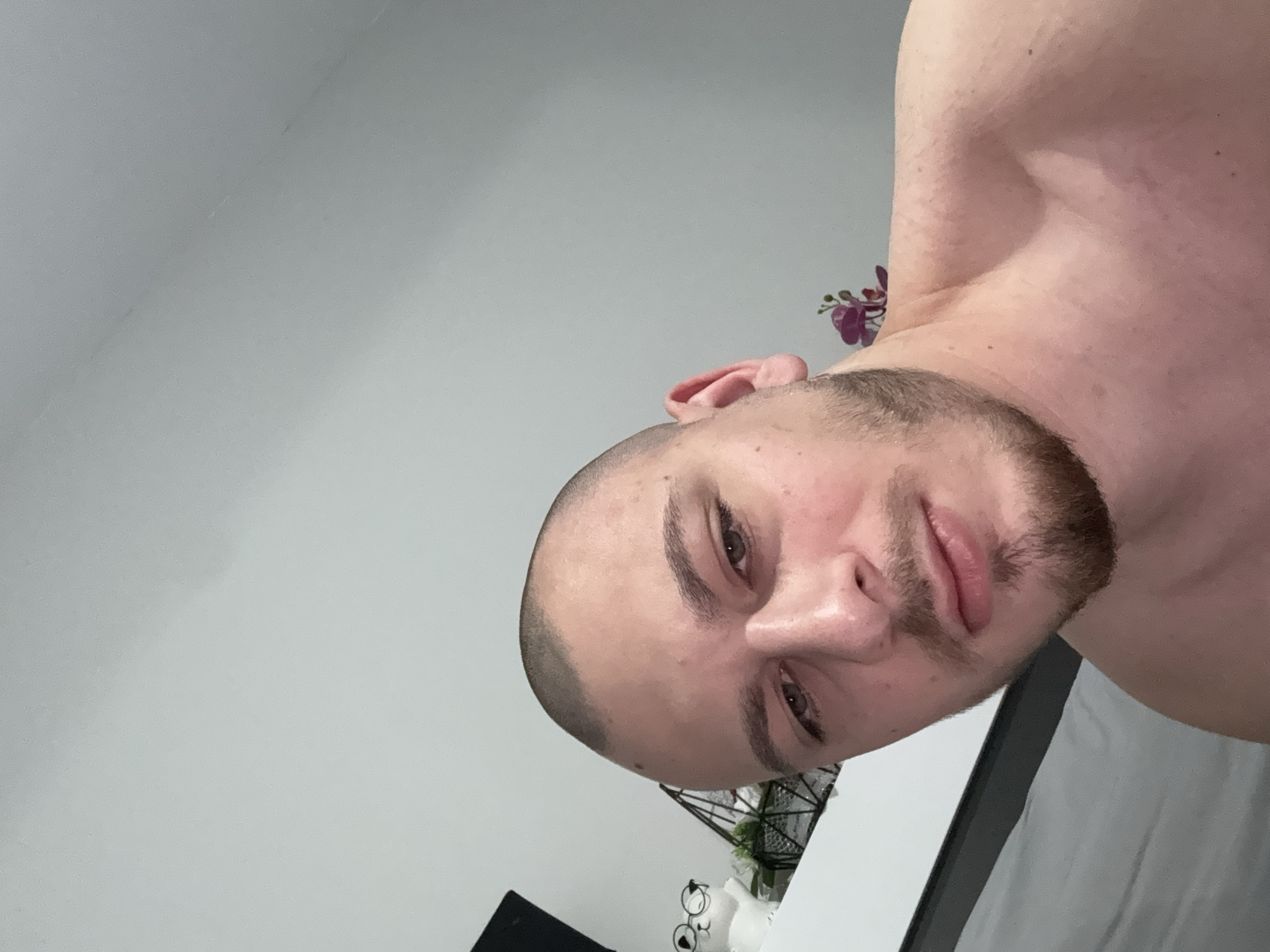 A close-up, slightly off-kilter, portrait-oriented photograph shows a man with a shaved head and a short beard. The man's face is the primary focus, with his eyes, nose, and mouth clearly visible. His skin tone is fair, and there are subtle facial features. The background is blurred and indistinct, but a white wall and a few decorative objects can be discerned. The lighting is soft and diffused. The photograph appears to have been taken indoors, with the man holding the camera at an angle. The photograph doesn't contain any information to conclude the location in the city of Stara Zagora, Bulgaria.