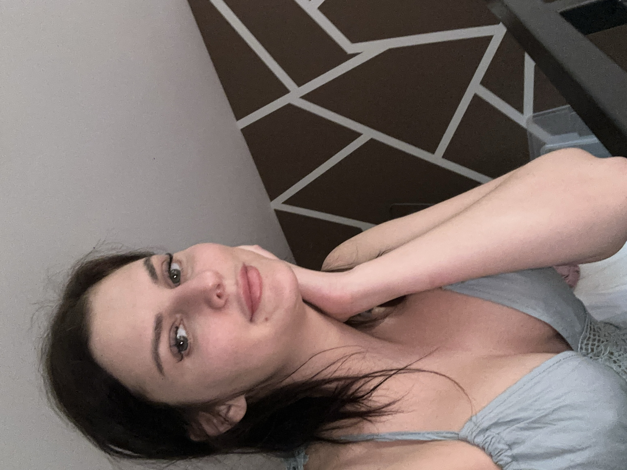 A medium shot captured from a slightly high angle shows a young adult Caucasian woman with medium-length dark brown hair, a light complexion, and green eyes. She is leaning on her hand, with her arm bent and resting on her cheek. Her head is slightly tilted, and she is looking directly at the camera with a gentle smile. She is wearing a light blue camisole with thin straps and lace trim. The background features a brown wall with a geometric pattern of white lines. The lighting is soft, and the overall atmosphere appears casual and intimate. The scene is devoid of any recognizable urban landmarks or specific environmental context that would indicate a location within a city.