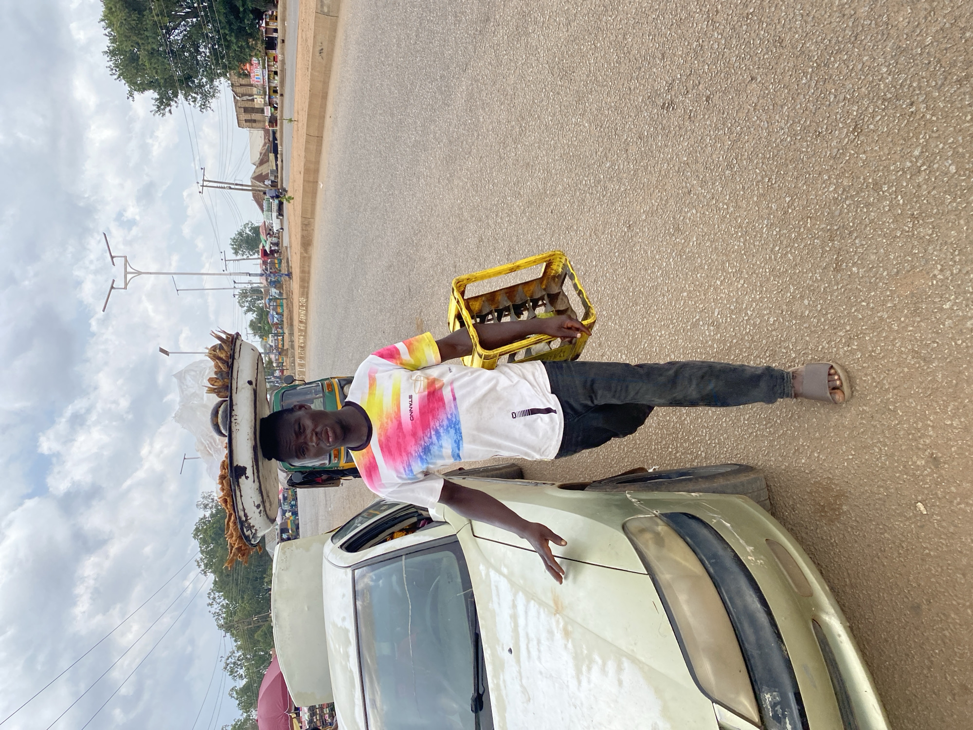 A young man, likely in his late teens or early twenties, stands on a dusty road in Bukuru, Nigeria. He is dressed in a white t-shirt with a colorful, abstract print and dark trousers. He is barefoot, wearing only sandals. On his head, he balances a large, white, oval-shaped platter piled high with what appears to be fried plantains or a similar snack. He is also holding a yellow plastic crate filled with bottles, possibly beverages, in his right hand.
Behind him, a light-colored car with its hood up is parked on the side of the road. The car appears to be older and shows signs of wear. Further in the background, the road continues, with a line of market stalls and shops visible on the left side. People are milling about, and various vehicles, including a green auto-rickshaw, are present. Electrical poles and wires crisscross the scene, indicating a built-up area.
The sky is filled with large, white clouds against a pale blue backdrop, suggesting it might be daytime, perhaps mid-morning or afternoon. The overall impression is a busy street scene in a Nigerian town, where street vendors are a common sight. The young man appears to be working, transporting goods for sale. His facial expression is neutral, but he seems aware of the camera. There is no visible text in the image.