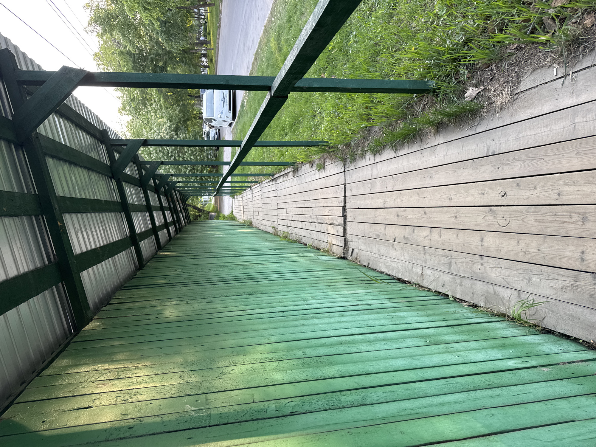 The image depicts a green-painted wooden walkway or ramp with a protective metal roof on one side and a wooden fence/wall on the other, located in the city of Khabarovsk, Russia. The walkway appears to lead upwards. On the right side, beyond the wooden wall, is grass. In the distance, cars are visible on a road. The day appears to be sunny. The structure of the roof and walkway is made of wood.