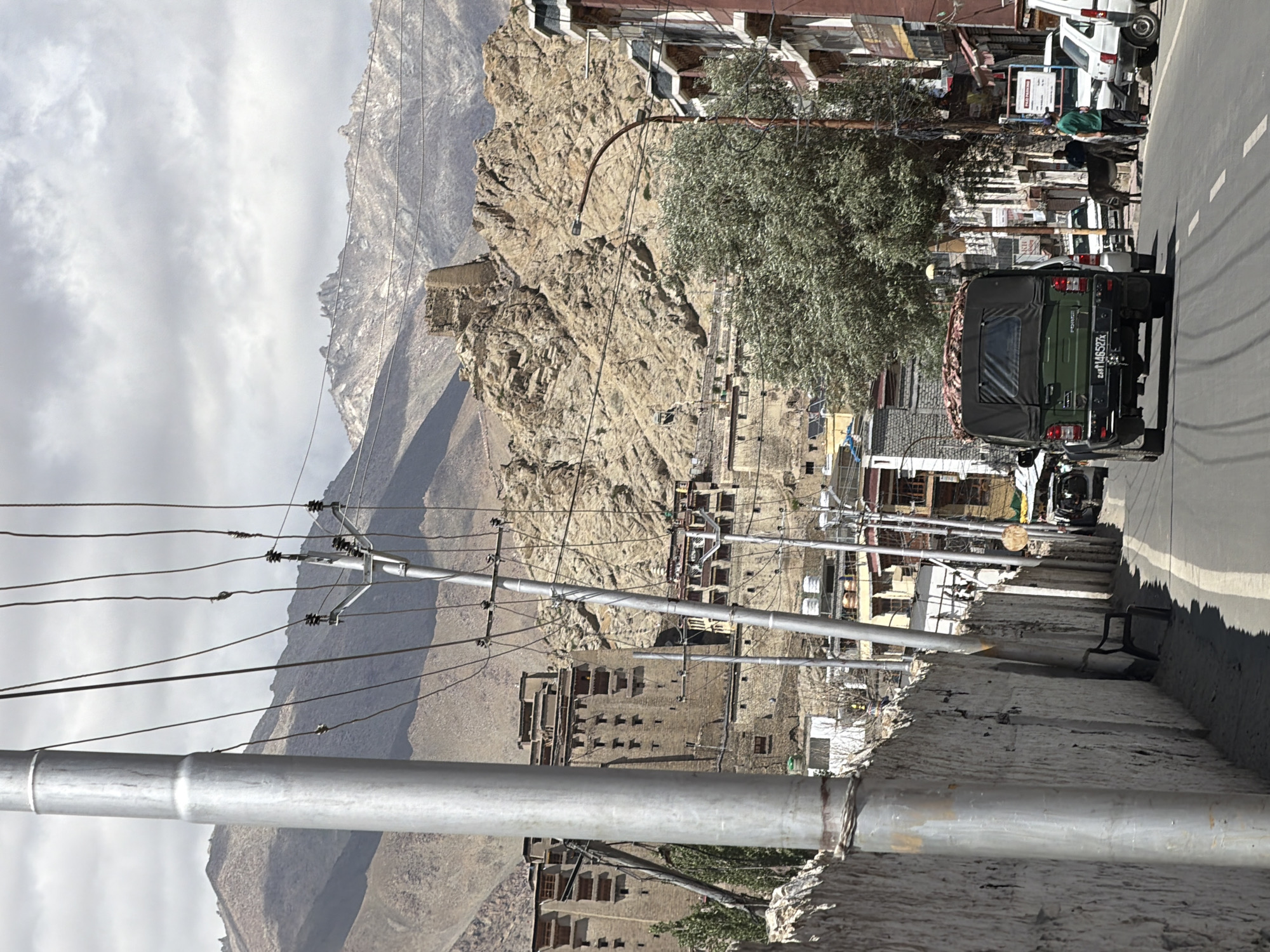 The image captures a street scene in Leh, India, under a cloudy sky. In the foreground, a large metallic pole stands to the left, with a white, textured wall running alongside the street. Utility poles and wires crisscross the frame, indicating an urban or semi-urban setting.
A dark green military-style vehicle with a covered cargo area is driving down the road, its license plate reading "ZA8 T 146527X". Beside the vehicle, a donkey is present, being led by a person in a green shirt. Several other vehicles, including white cars, are parked or moving along the street.
The background reveals the distinctive landscape of Leh, with traditional buildings constructed from stone and mud, appearing to be integrated into the rocky hillsides. These structures, reminiscent of a historical fort or monastery, are nestled against the rugged, mountainous terrain that dominates the backdrop. The overall atmosphere suggests a blend of daily life and the unique architectural and natural environment characteristic of Leh.