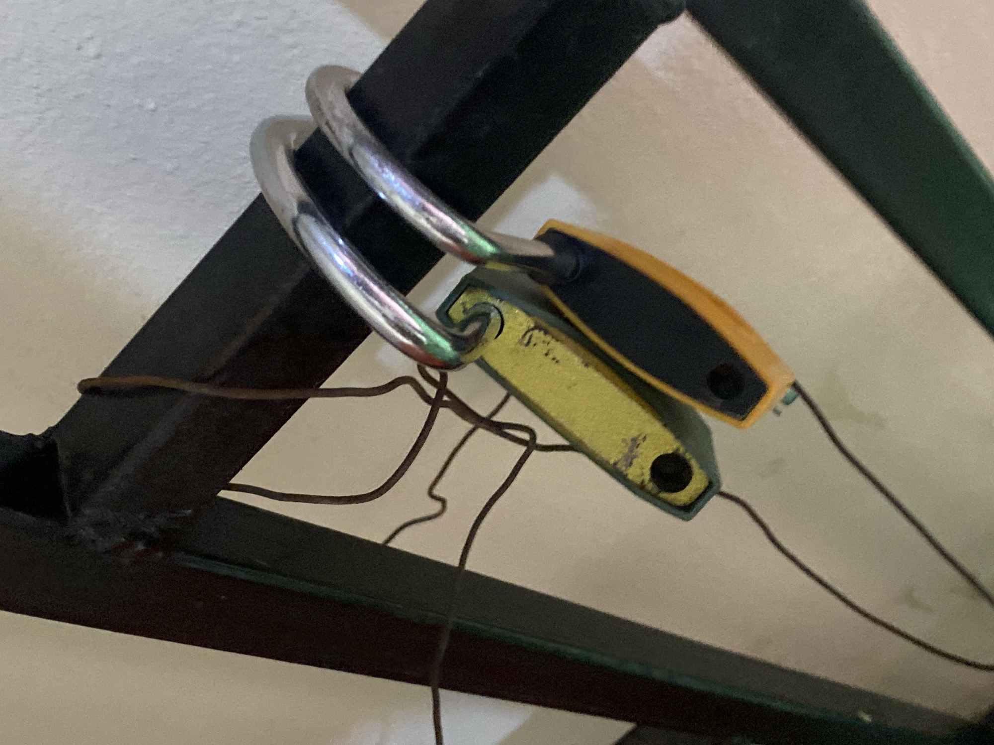 In Jauro Yinu, Nigeria, a close-up shot focuses on two yellow and black electronic tags attached to a metal frame by a silver padlock. The tags, one predominantly yellow with black accents and the other black with yellow accents, are connected to wires that hang loosely. The metal frame appears to be part of a larger structure, possibly furniture or a gate, with a rough, dark finish. The background is a plain, light-colored wall. The lighting is artificial and casts subtle reflections on the metal padlock.
