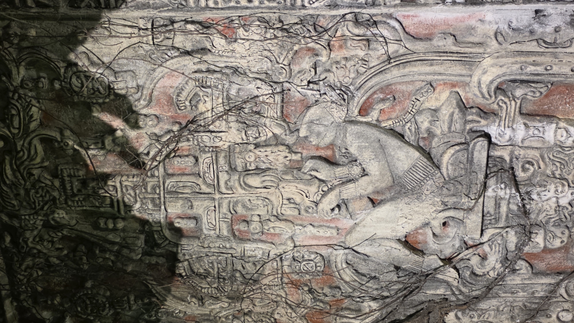 The image depicts a bas-relief carving, possibly of Mayan origin, with a figure kneeling and interacting with glyphs. The carving is rendered in stone, exhibiting wear and discoloration. Traces of red pigment remain within the relief. The scene is obscured by vegetation and shadows. The city of origin is unknown.