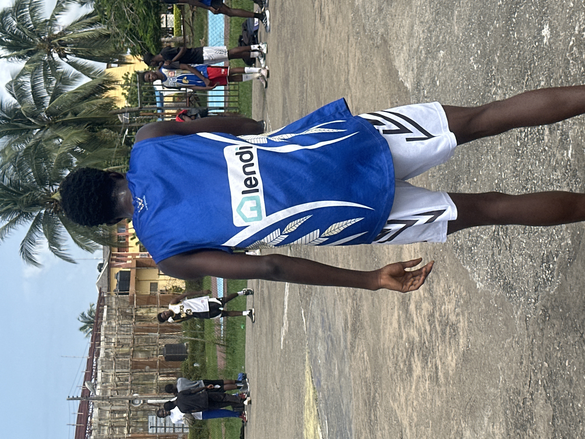 An African male, facing away from the camera, stands on a concrete basketball court. He is wearing a blue basketball jersey with white trim and a logo that reads "lendo," and white basketball shorts with black accents. His dark hair is cut short. In the background, other individuals are present on the court, some wearing basketball attire. The court is outdoors and appears to be in a city setting, possibly Ajara, Nigeria, given the presence of palm trees and the architectural style of the buildings in the distance, which include what appears to be a partially constructed building with scaffolding. The sky is overcast.
