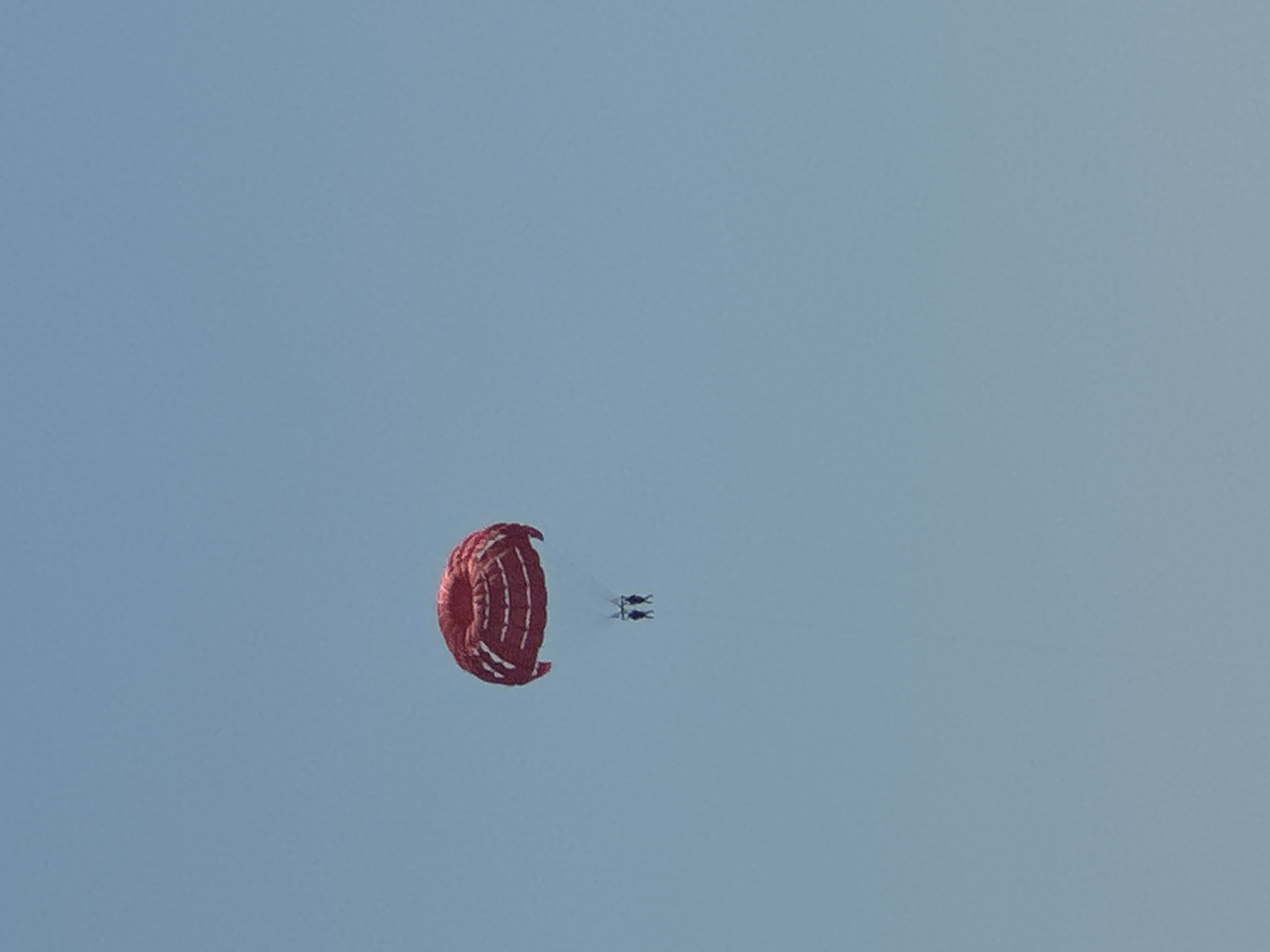 Two people are parasailing high in the clear, pale blue sky over Budva, Montenegro. A large, red parachute, possibly a parasail, is open above them, billowing slightly. The two individuals are suspended beneath the parachute by lines, appearing as dark figures against the sky. The scene is serene and focuses on the activity of parasailing against a vast expanse of sky, suggesting a leisurely or recreational moment.
