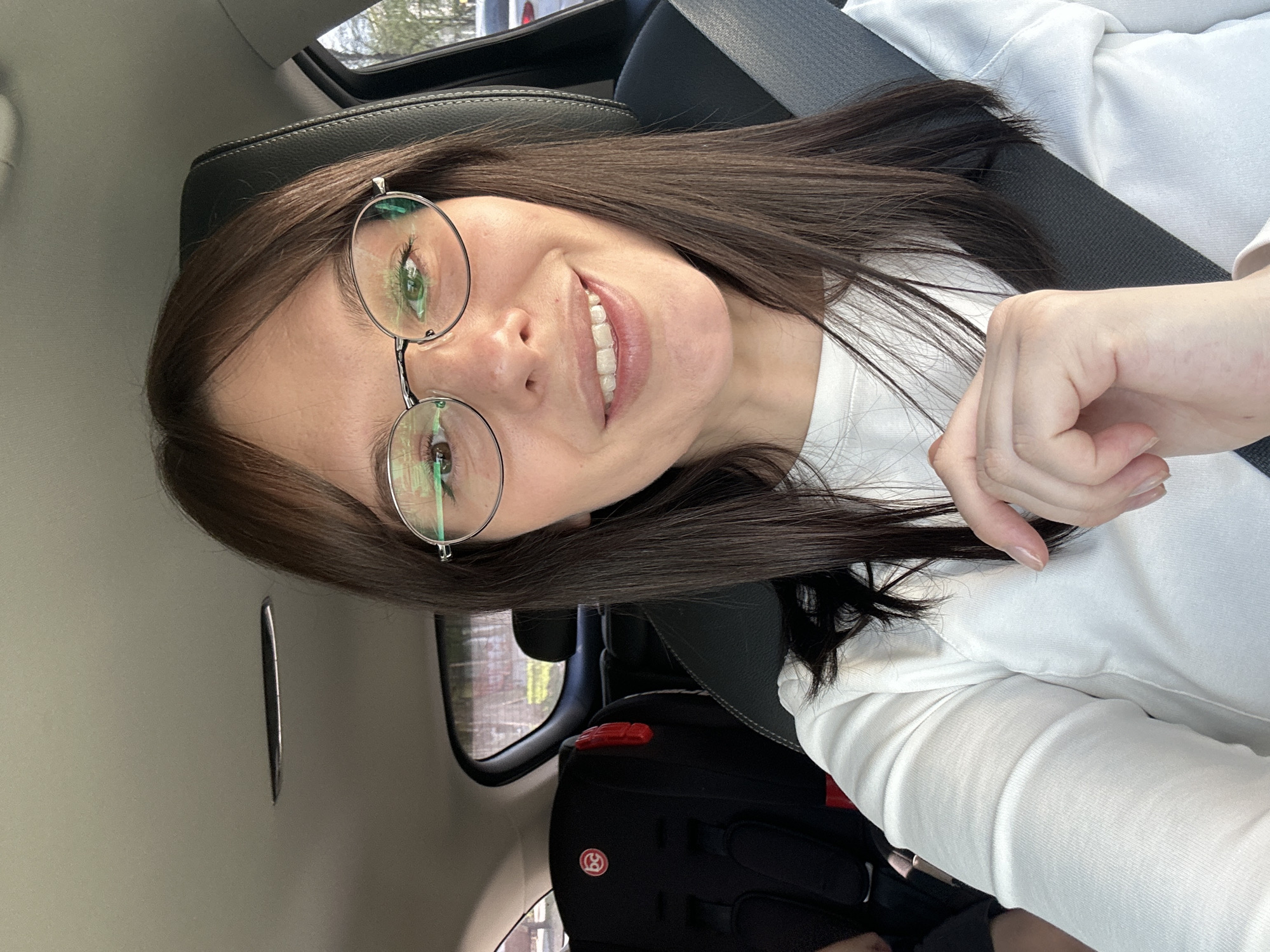 A woman with long brown hair, wearing round glasses and a white shirt, is in a car. She is wearing a seatbelt. A child's car seat is behind her. The location appears to be inside a vehicle in an unidentified location; no definitive elements indicate the city of Chaikovsky, Russia.