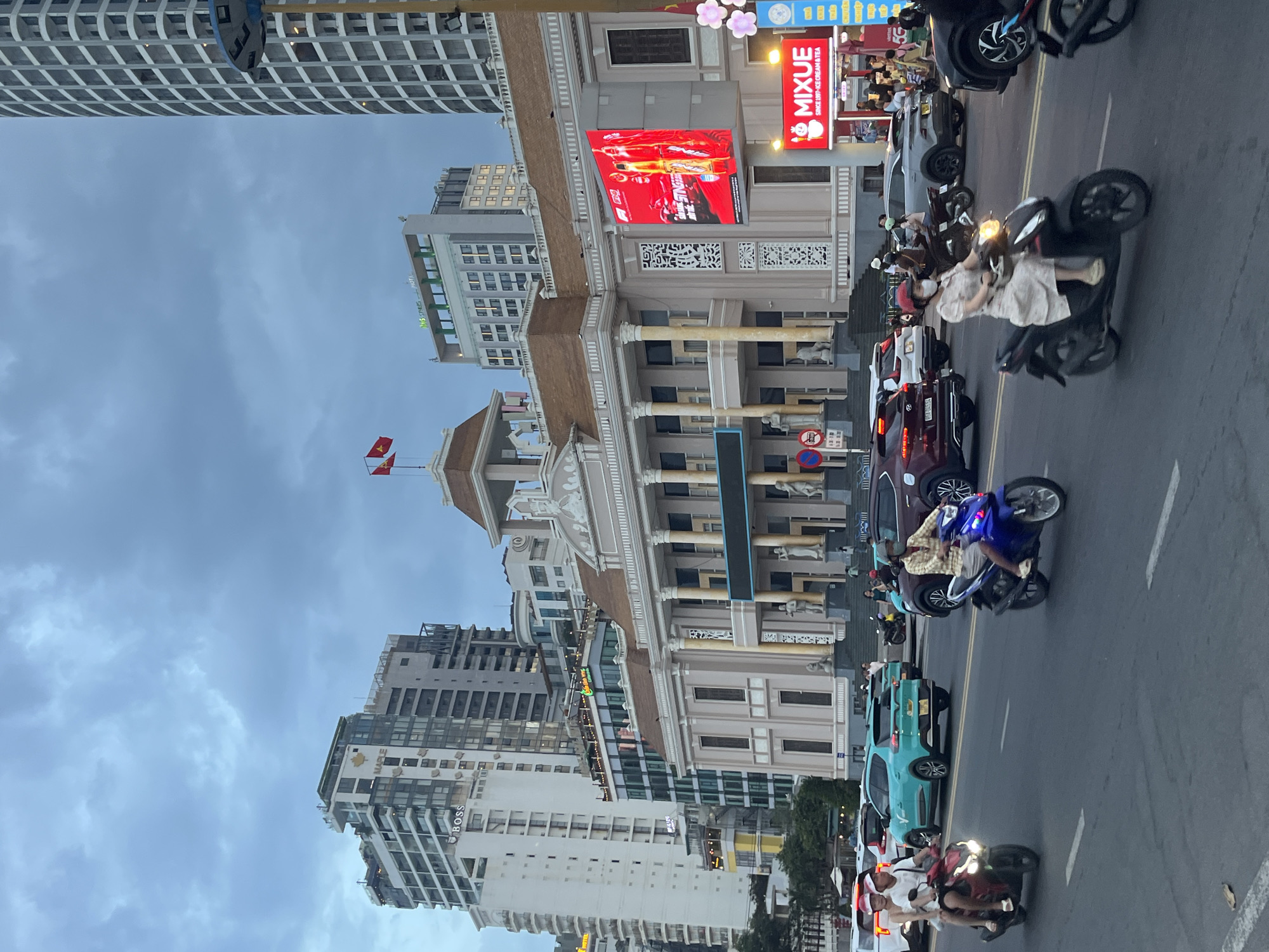This is a street-level, eye-level, vertical, full shot taken at dusk in Nha Trang, Vietnam, focusing on the Ho Chi Minh City Museum of Fine Arts building. The sky is overcast with dark clouds. A multi-story building with classical architectural elements, including columns and a pediment, dominates the central portion of the frame. A large digital billboard displaying a Formula 1 advertisement is mounted on the side of this building. To the right, a smaller red sign with the text "MIXUE SINCE 1997 ICE CREAM & TEA" is visible.
In the foreground, a street is filled with traffic. Several motorcycles are in motion. On the left, a red motorcycle carries two people, the passenger wearing a red Santa hat. Next to it, a light blue SUV is in the lane. Further back, a dark red SUV is seen. On the right side of the frame, a woman in a light-colored dress rides a black scooter, wearing a face mask. Another scooter is visible to her right. Beyond the vehicles, pedestrians can be seen walking.
In the background, numerous modern high-rise buildings of varying heights and architectural styles are visible, with some featuring illuminated windows. Two Vietnamese flags are flying from a rooftop on the left side of the central building. The overall scene depicts a busy urban street during the evening, with a mix of historical and modern architecture and active street life.