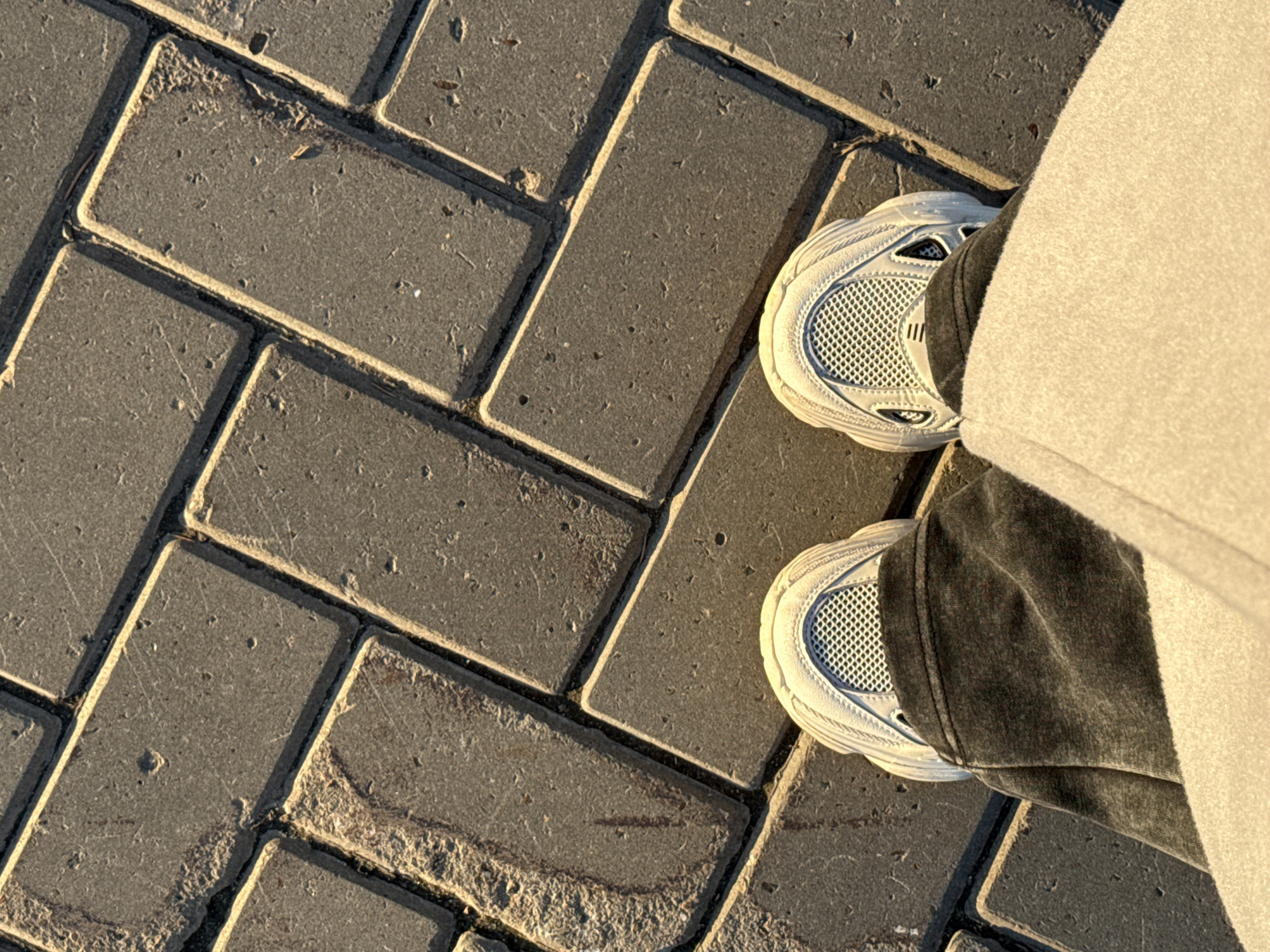 A top-down shot captures a person standing on a paved surface constructed from grey rectangular bricks arranged in a herringbone pattern. The right side of the frame shows the person's feet, clad in white sneakers with a mesh pattern on the upper part of the shoe. The person is also wearing dark grey or black pants. A light-colored coat or other garment appears to be draped or worn on the same side, partially obscuring the shoe and pants. The image has a muted tone, and a shadow is cast across the bricks, suggesting the photo was taken during the day, possibly near dawn or dusk. The scene offers no clear indication that the location is Sviyazhsk, Russia.